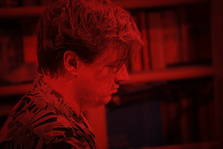Man met lichtbruine haar en een bloemenprint shirt, zijne ogen dicht en lijkt zich te concentreren, op de achtergrond een boekenkast in een kamer met een rode tint.