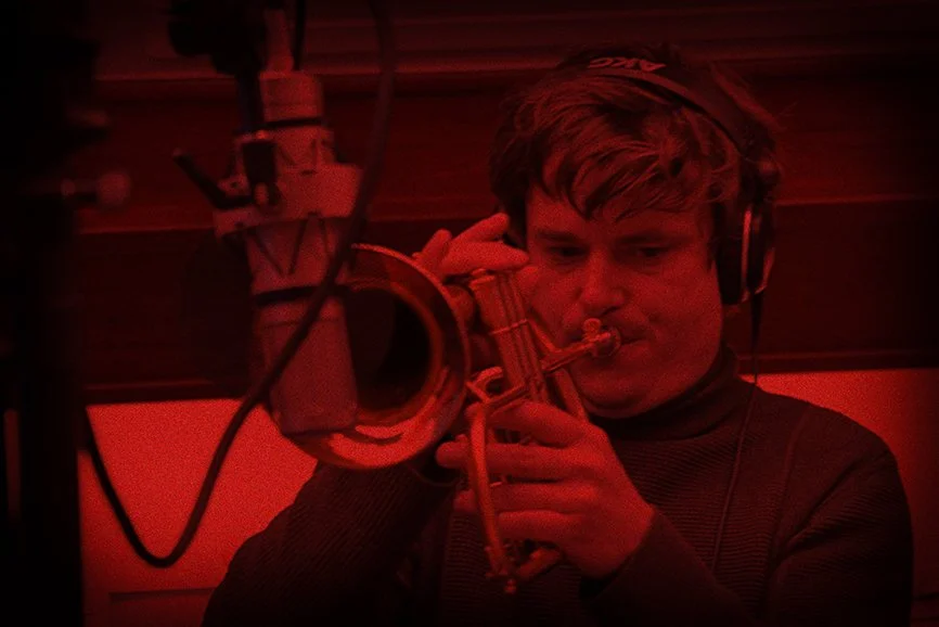 Muzikant die trompet speelt in een opnamestudio, verlicht door rood licht, met koptelefoon op.
