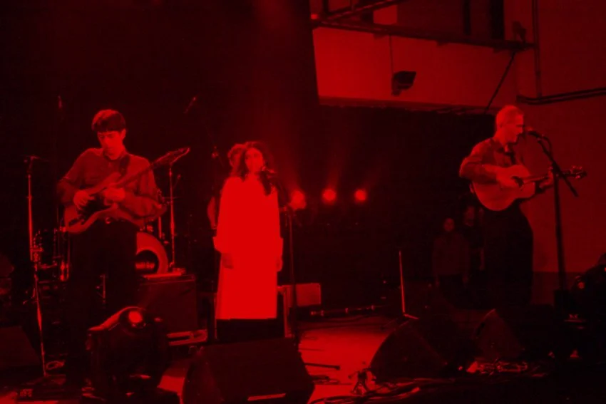Muziekband op het podium tijdens een live optreden, met drie muzikanten onder rood verlichte omstandigheden.