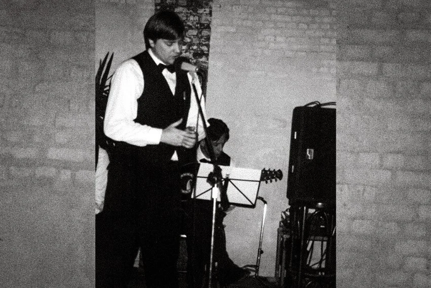 Een man die zingt en gitaar speelt tijdens een optreden, met een andere muzikant op de achtergrond, in een ruimte met een bakstenen muur.