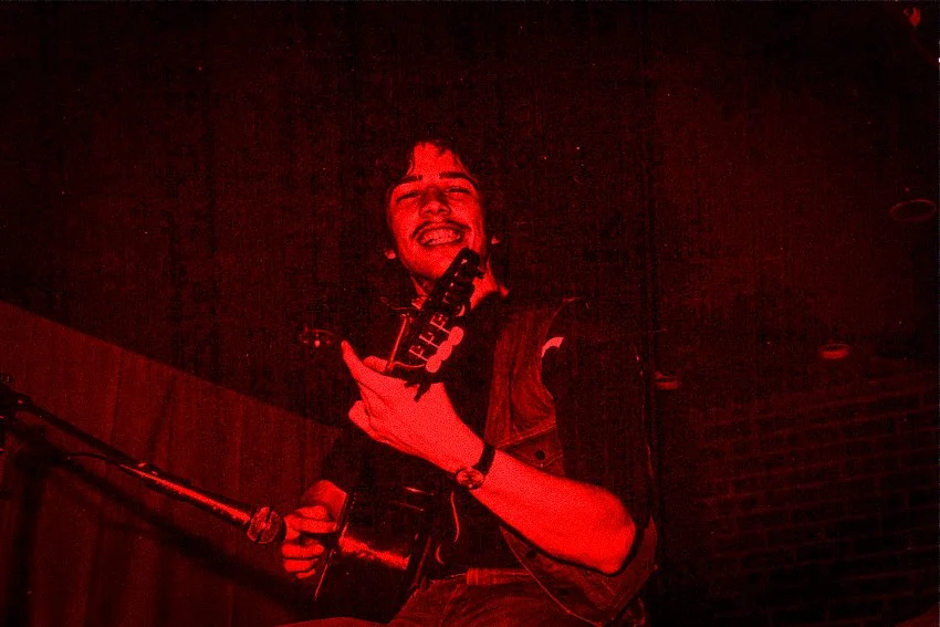 Een jonge man met een glimlach, die gitaar speelt op een podium onder rood licht.