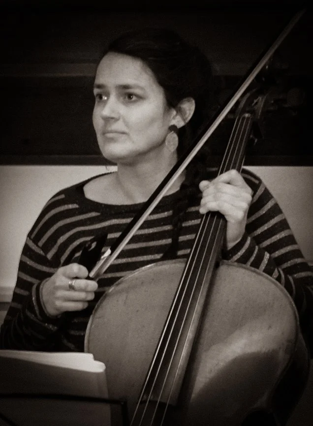 Een vrouw die een cello bespeelt, zie haar met een strijkstok in haar rechterhand en haar linkerhand op de vingers van de cello, in een donkere ruimte.