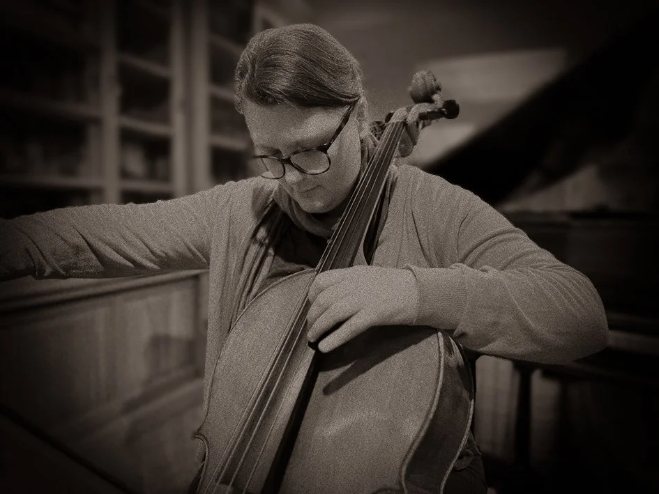 Een vrouw met bril die op een piano speelt en een cello vasthoudt, in een muziekkamer.