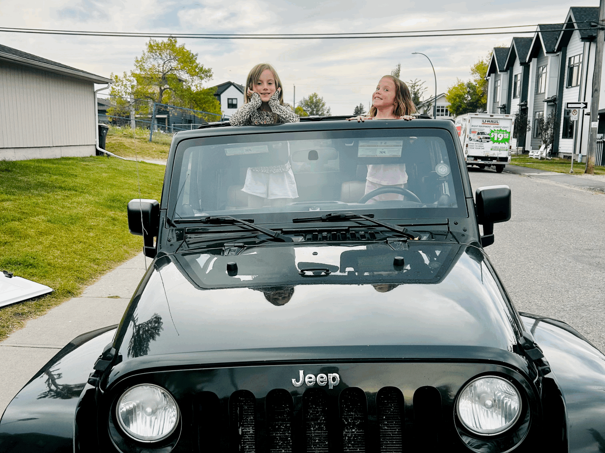 Two young girls sitting inside a black Jeep, with one girl standing and the other girl sitting, enjoying a ride on a suburban street during daytime.