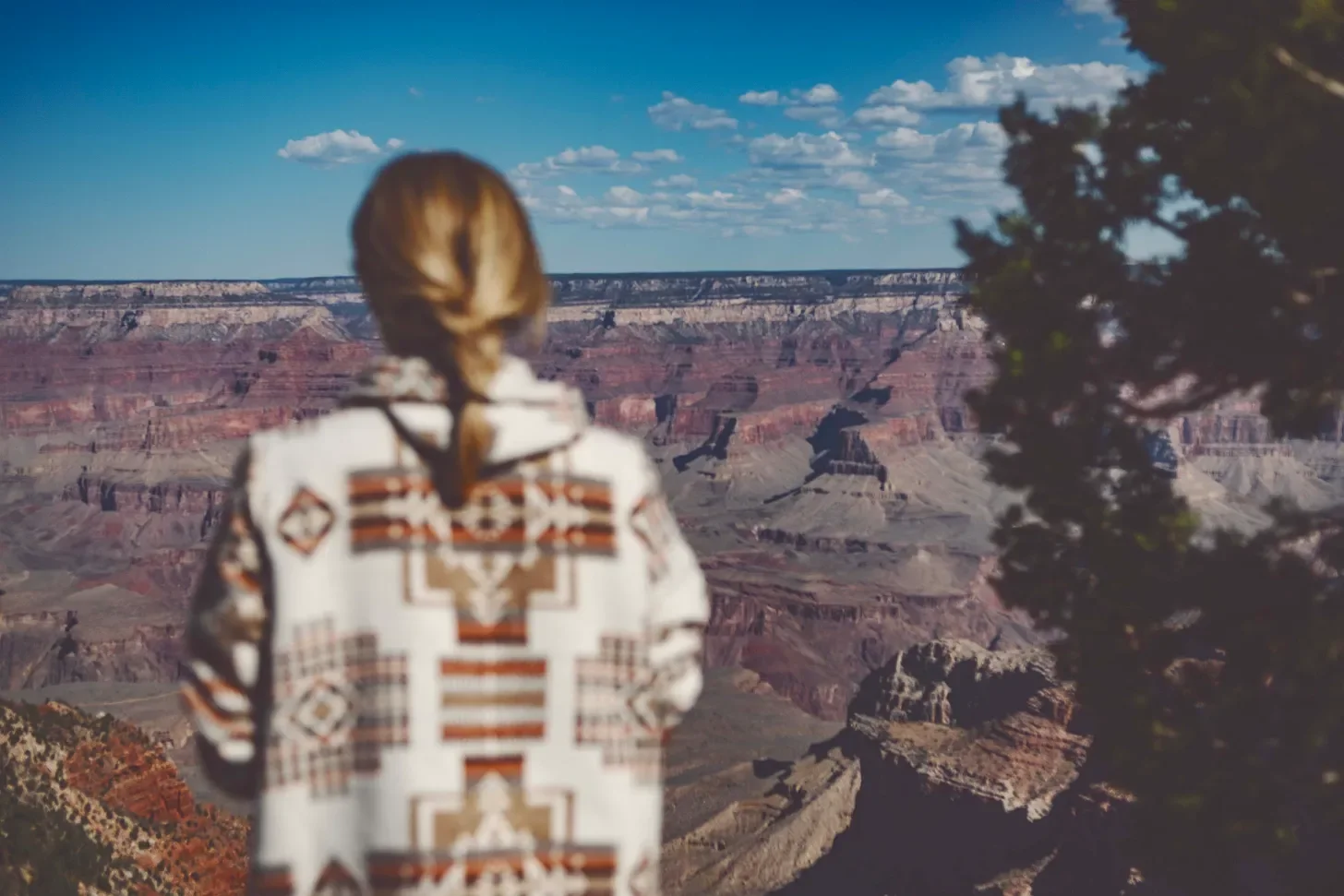 A woman with brown braided hair wearing a patterned jacket stands with her back to the camera, overlooking the Grand Canyon on a clear day with scattered clouds.