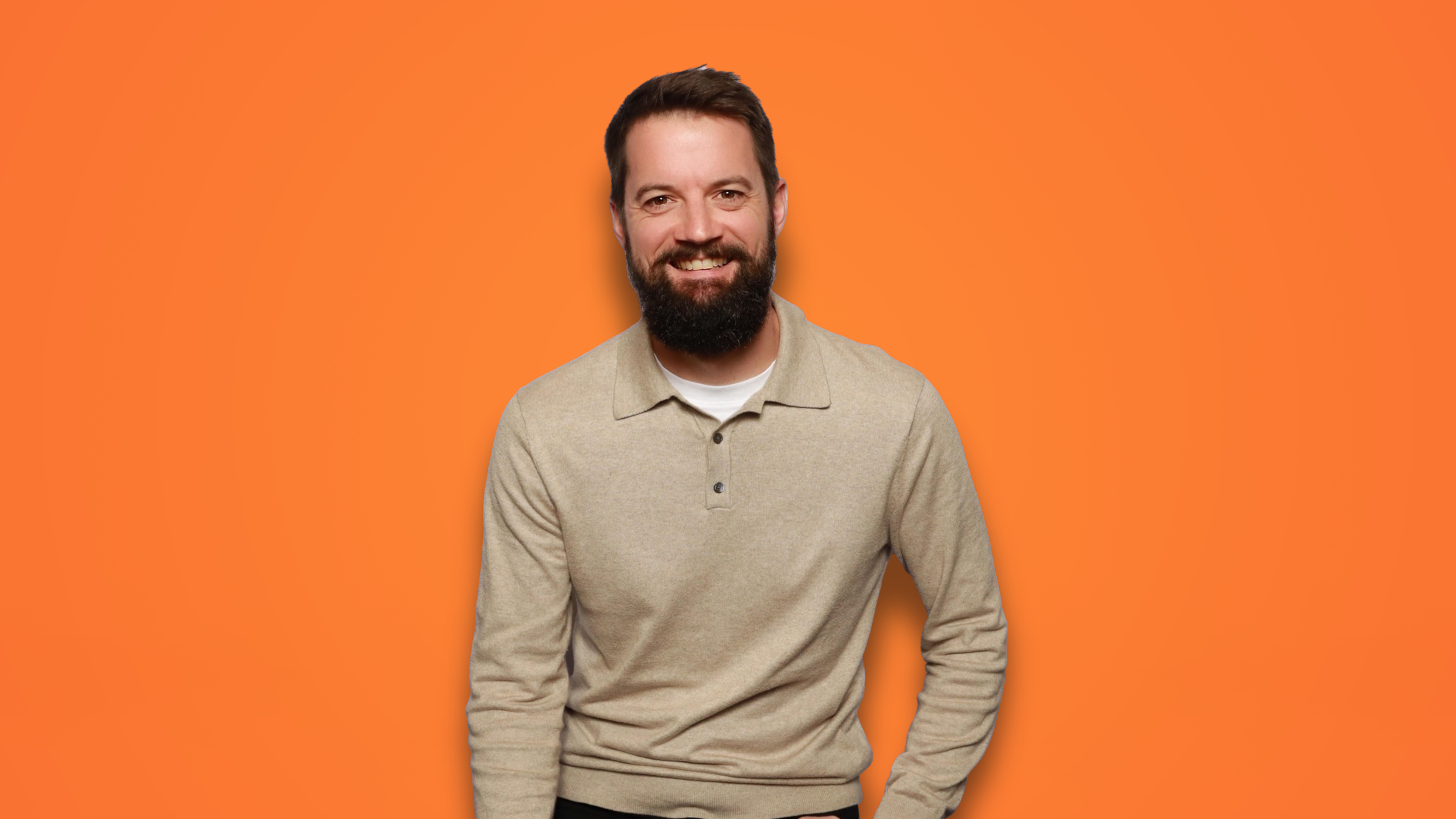A man with a beard and short brown hair smiling, wearing a beige long-sleeve polo shirt over a white shirt, against an orange background.