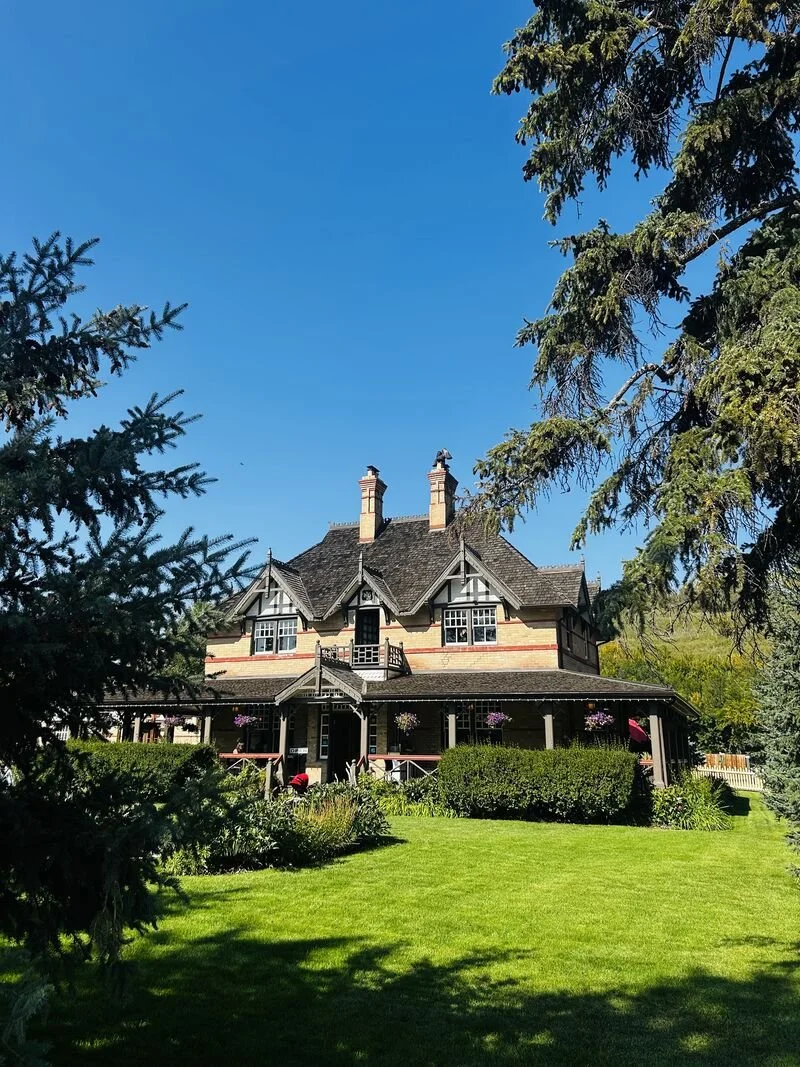 A large, historic house with a steep roof, multiple chimneys, and a wraparound porch, surrounded by trees and a well-maintained lawn under a clear blue sky.
