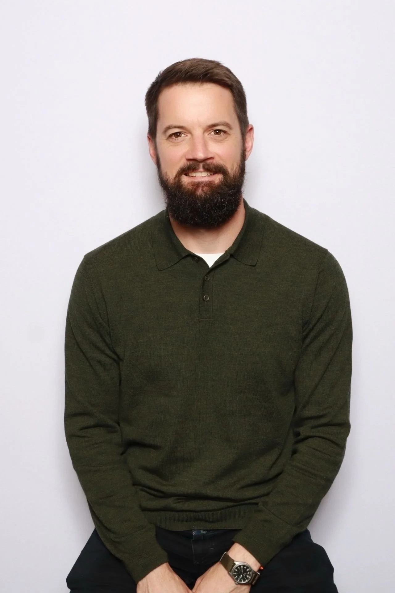A man with brown hair and a beard, wearing a dark green sweater, sitting against a plain white background.