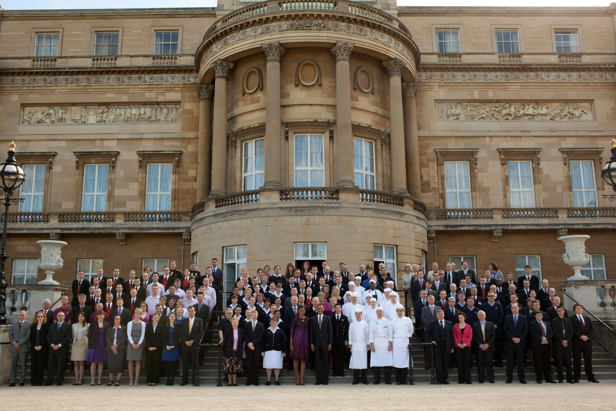 A large group of people, including chefs in white uniforms and hats, dressed in formal and professional attire, standing on the steps outside a historic building with classical architecture and decorative carvings.