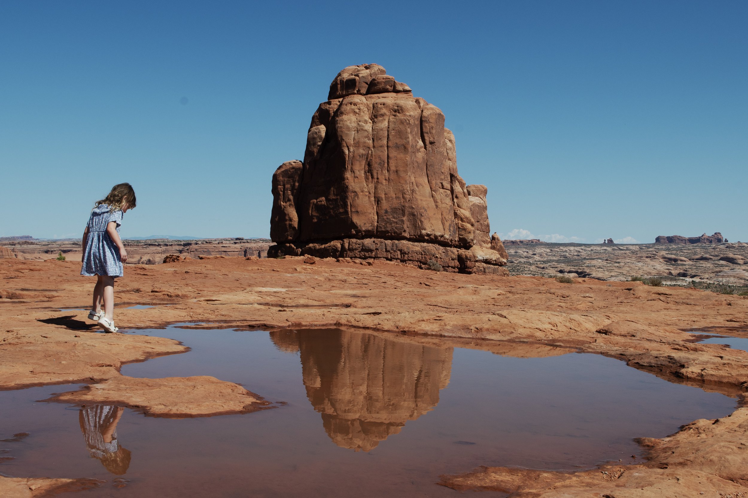 A young girl in a blue dress stands by a small water pool in a desert landscape, with a large red rock formation in the background and a clear blue sky.