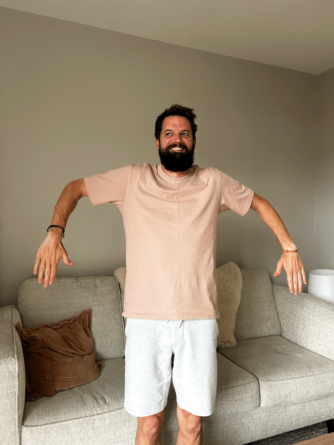 A man with a beard wearing a beige t-shirt and white shorts standing in a living room with his arms outstretched and holding a relaxed and happy expression.