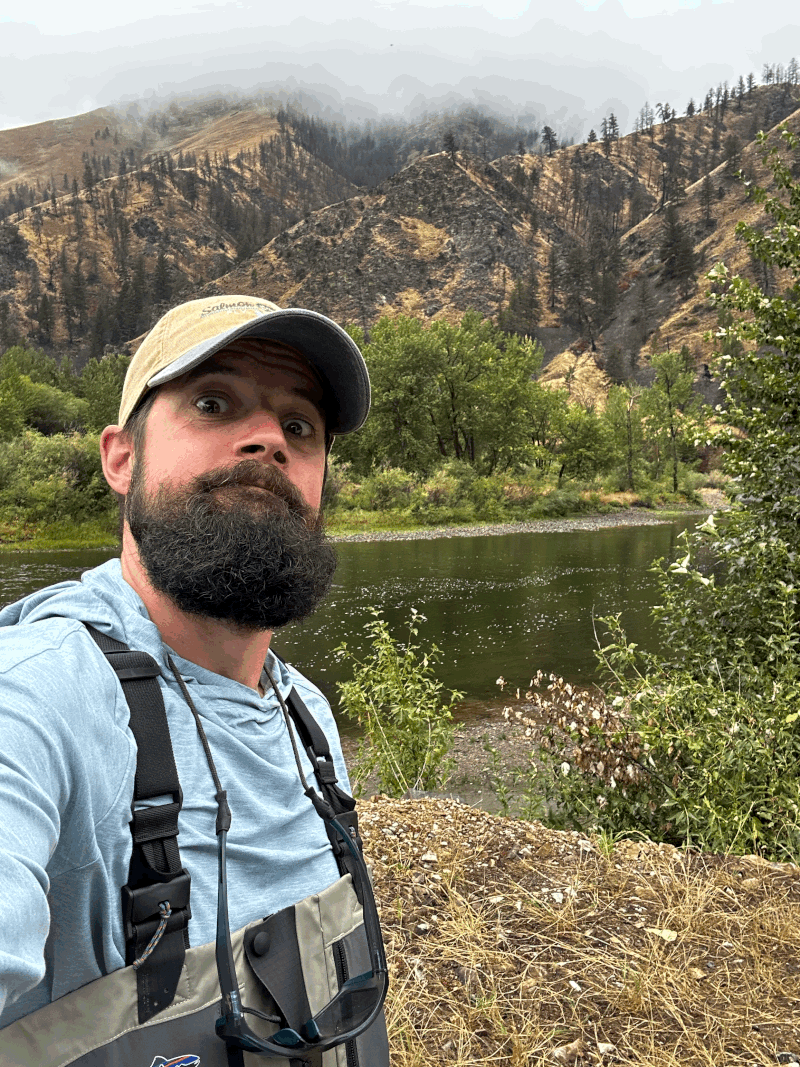A man with a beard and blue hoodie taking a selfie outdoors near a river with green trees and mountains in the background, some mountains appear to be cloudy.