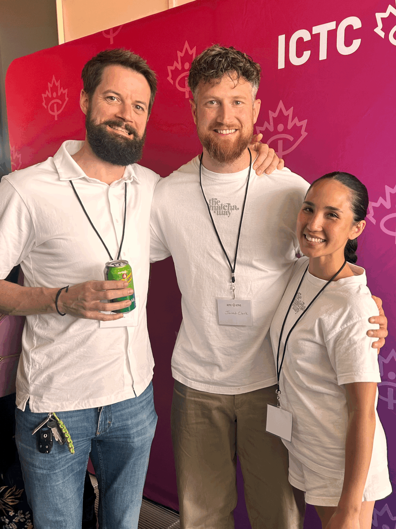 Three people standing together at the ICTC event, smiling and wearing white T-shirts with the event logo. The person on the left is holding a can of Sprite. They are in front of a pink and purple backdrop with the event name and logos.
