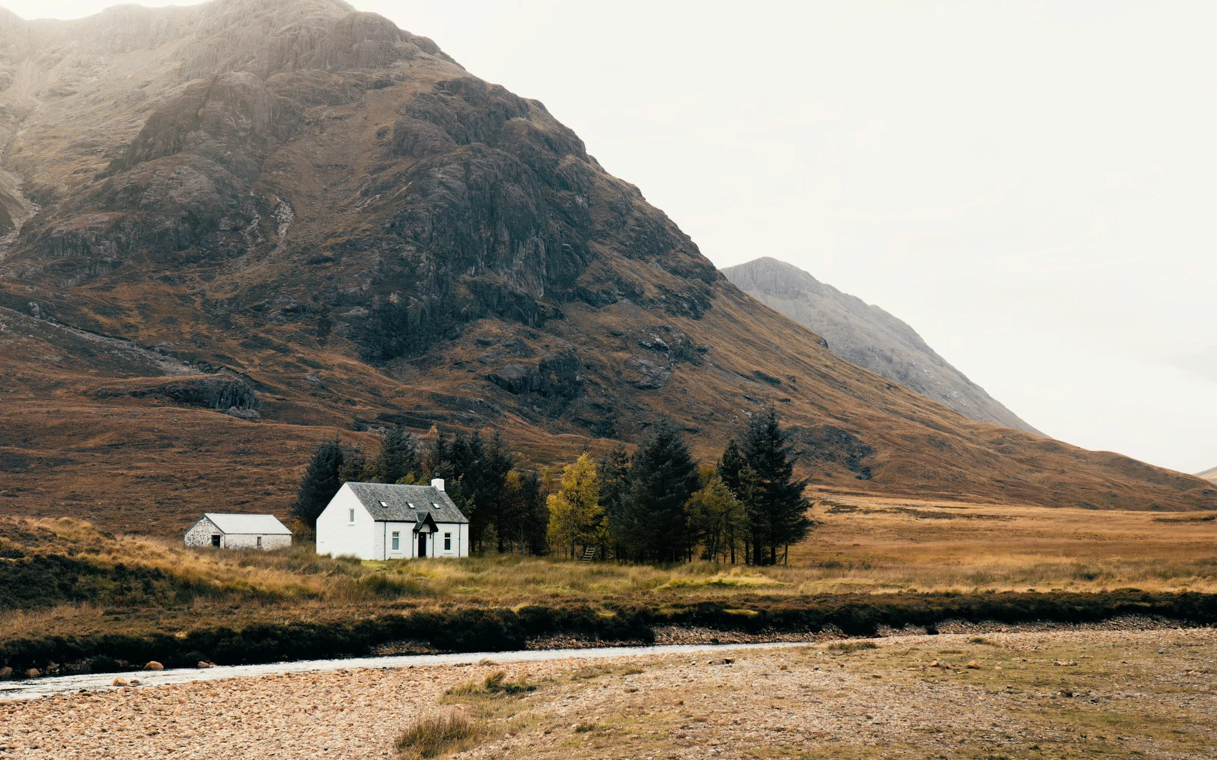 A white house with a smaller white shed nearby, surrounded by trees, set against a mountainous landscape with brown and green tones, a river running in the foreground.