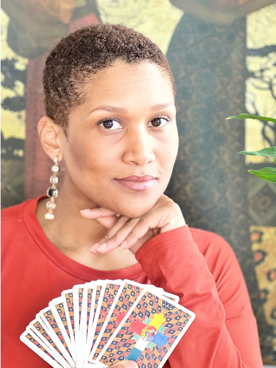 A woman with short curly hair, wearing a red top and decorative earrings, holds a fan of cards with geometric patterns and colorful illustrations, sitting in front of a nature-themed background.