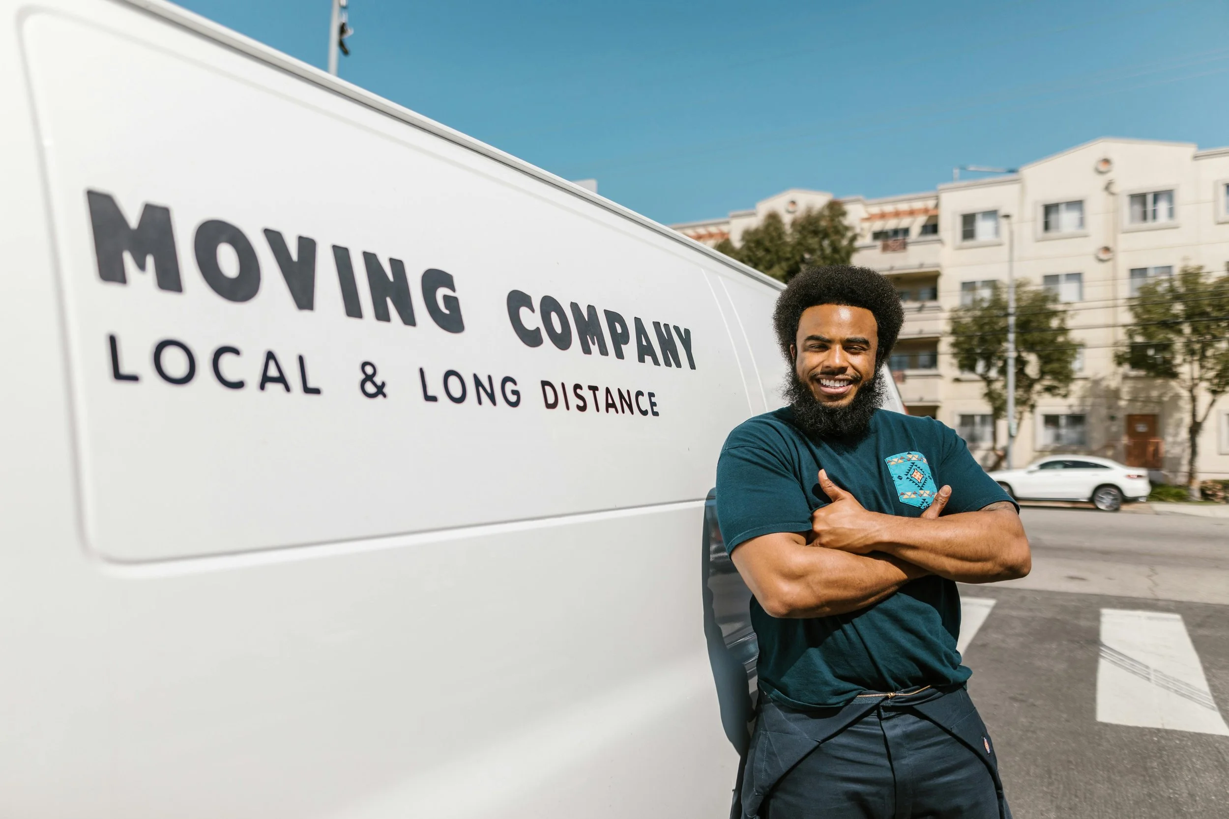 A man with a beard and curly hair smiling and crossing his arms in front of a moving company's truck on a city street.