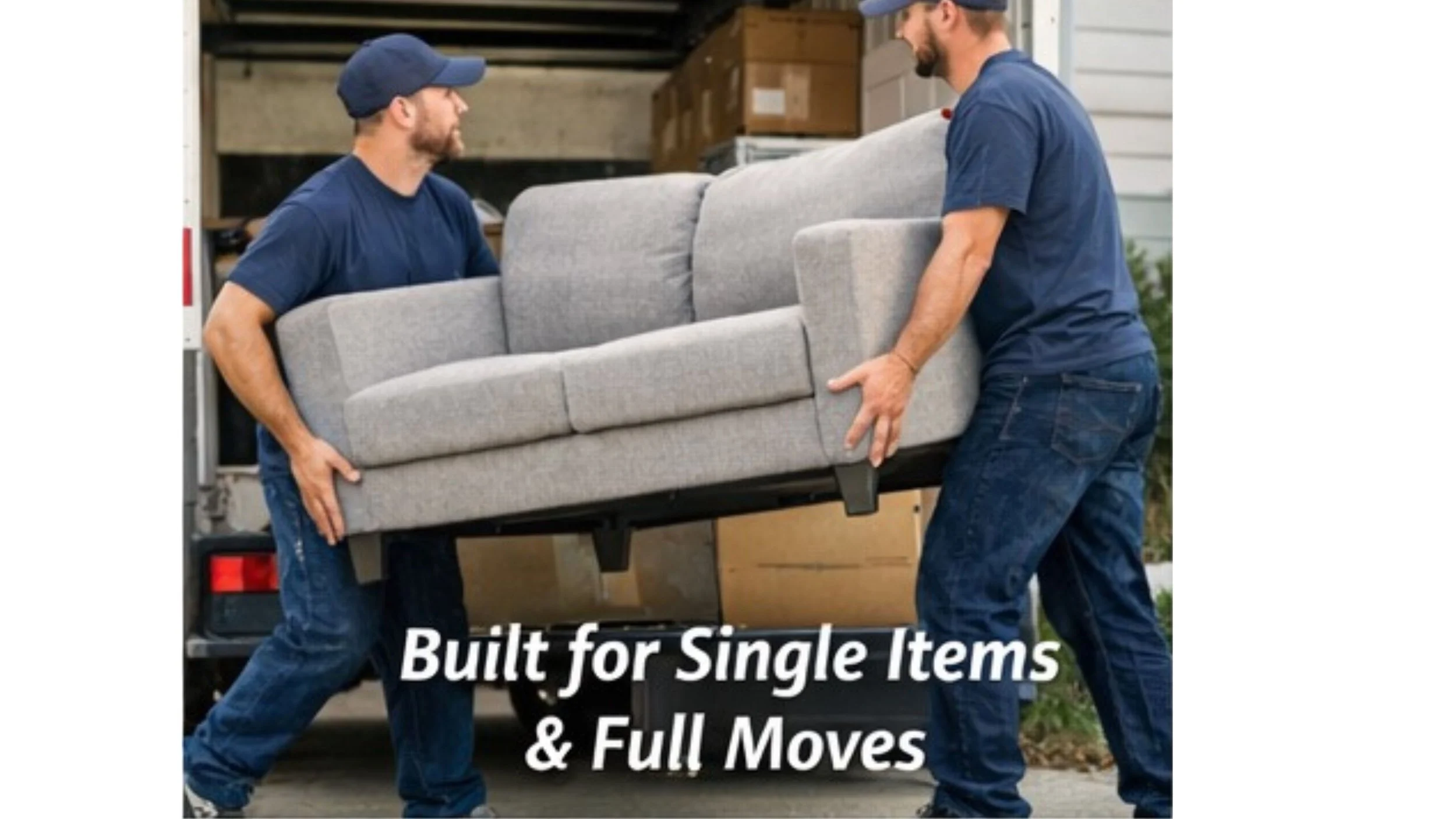 Two men are lifting a gray sofa into the back of a moving truck. They are wearing navy blue shirts and jeans, and are working together to move the furniture. The truck's open rear door shows boxes and other items stored inside. The text on the image reads: "Built for Single Items & Full Moves."