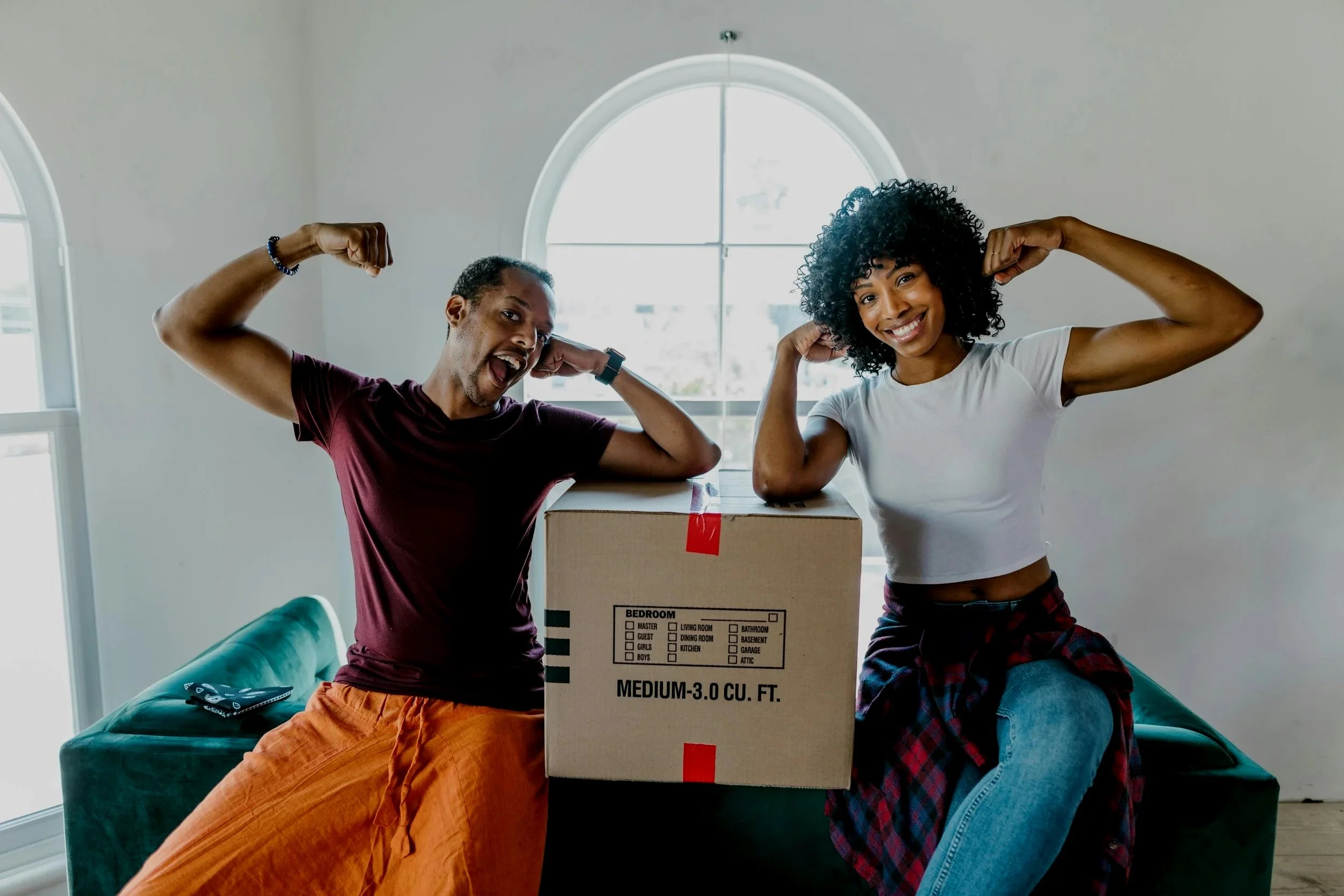 A happy man and woman flexing their muscles beside a cardboard box in a bright living room.