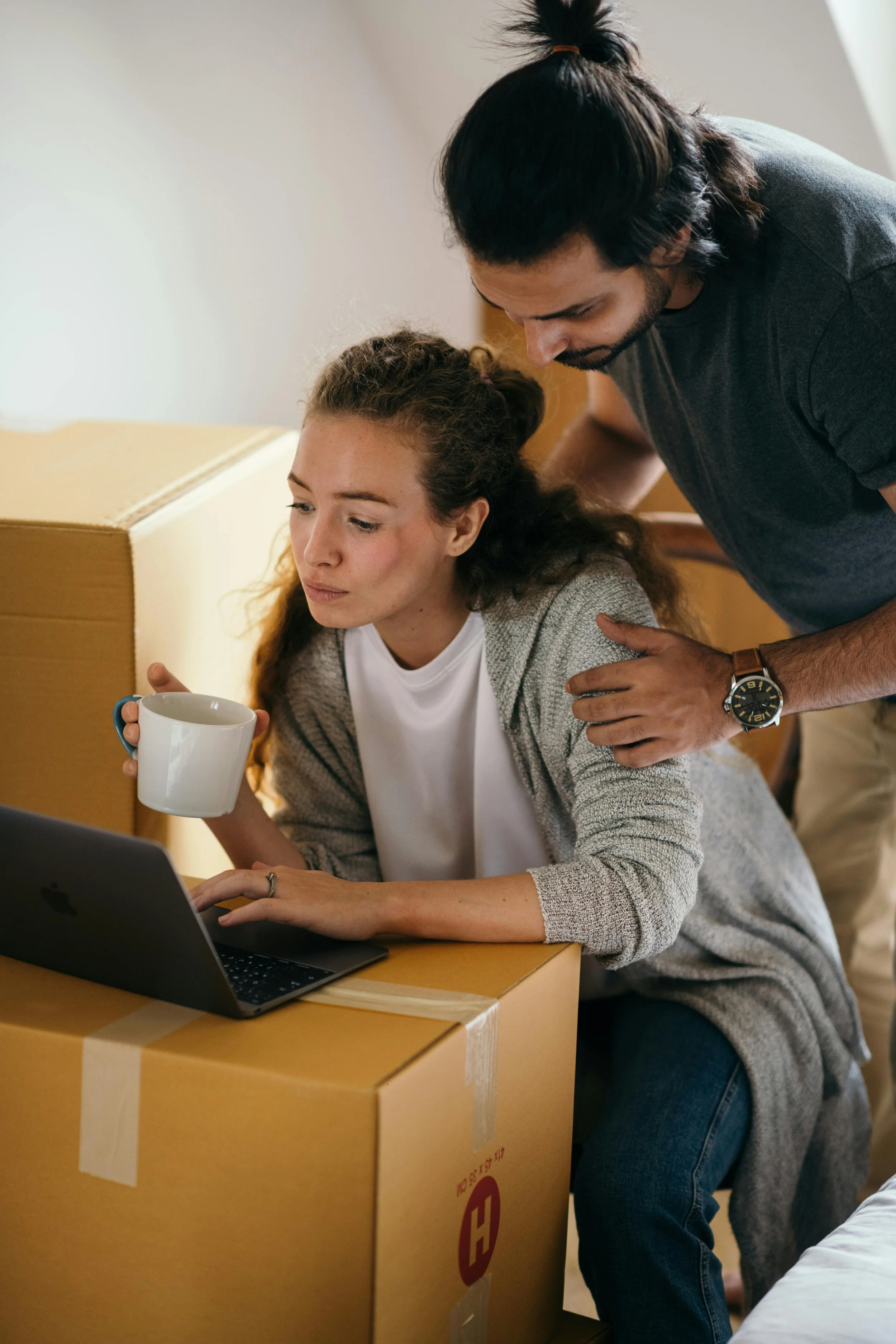 A woman sitting at a cardboard box with a laptop and a mug, while a man stands beside her, leaning over and looking at her screen.