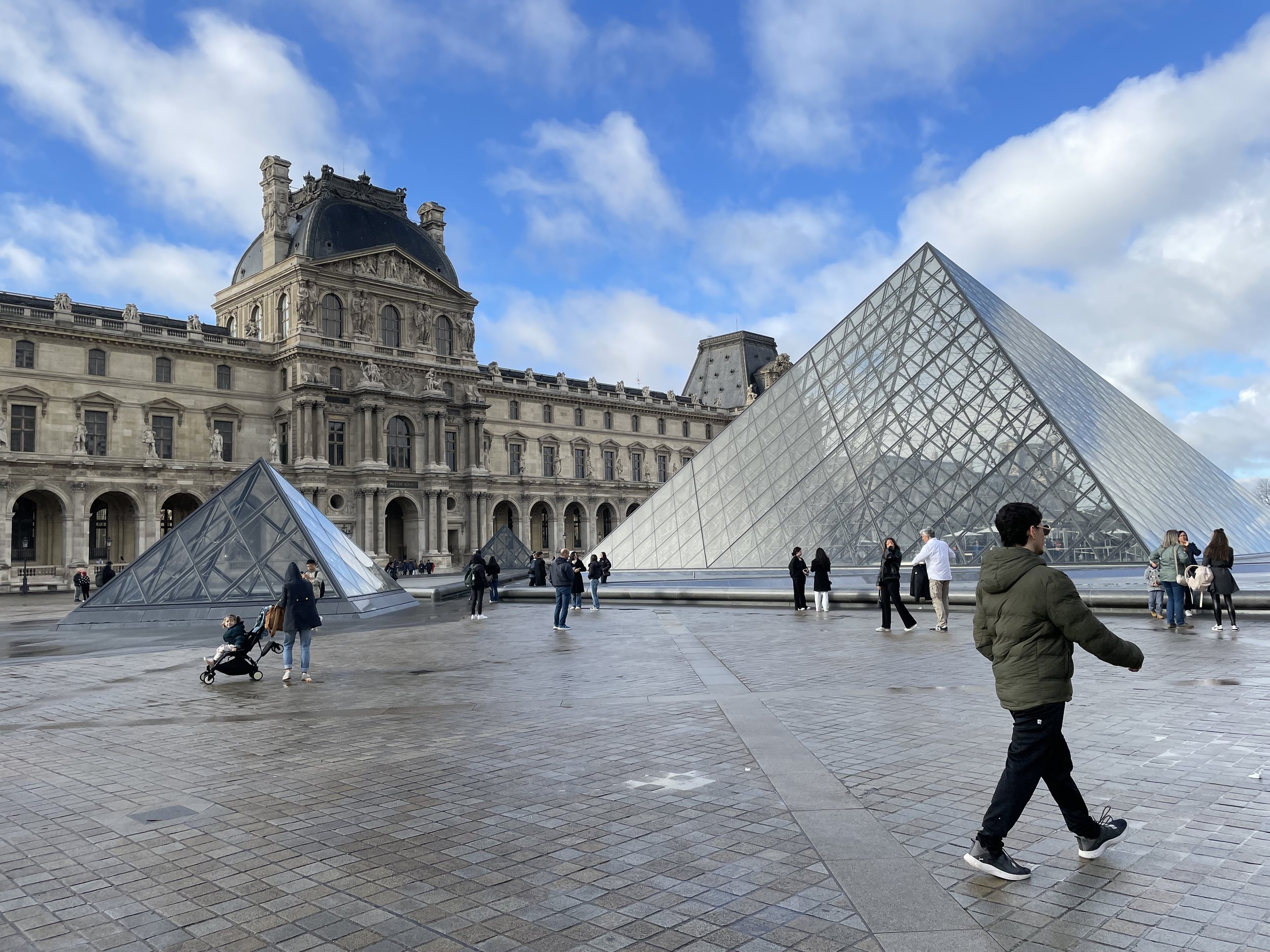 Vue du musée du Louvre à Paris avec ses pyramides de verre au premier plan et le bâtiment historique en arrière-plan sous un ciel bleu avec des nuages, des visiteurs se promènent autour.