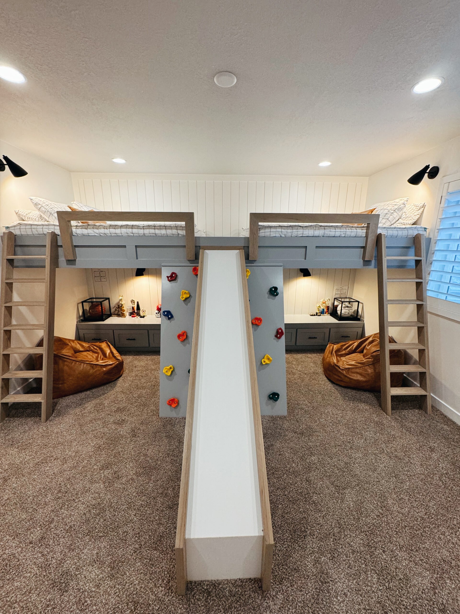 A children's two-story bedroom with a built-in play structure featuring a slide, climbing wall, and ladder, with beds on the upper level and bean bags below.