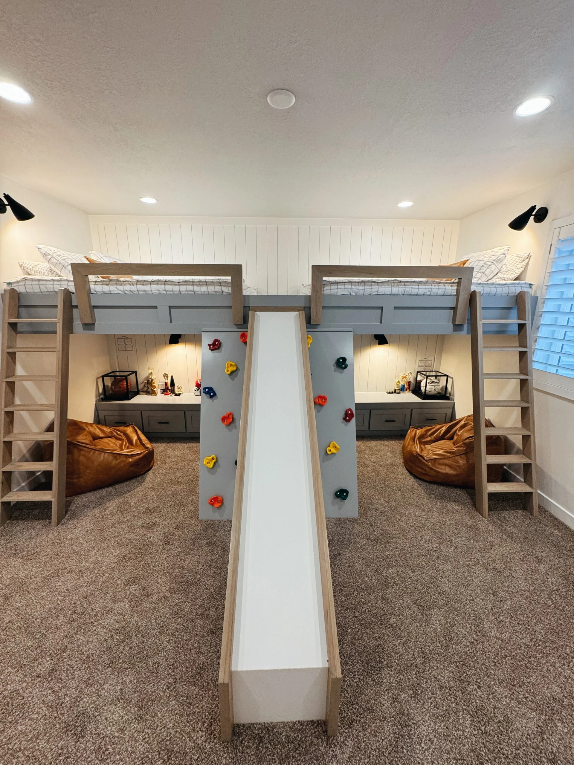 A children's two-story bedroom with a built-in play structure featuring a slide, climbing wall, and ladder, with beds on the upper level and bean bags below.