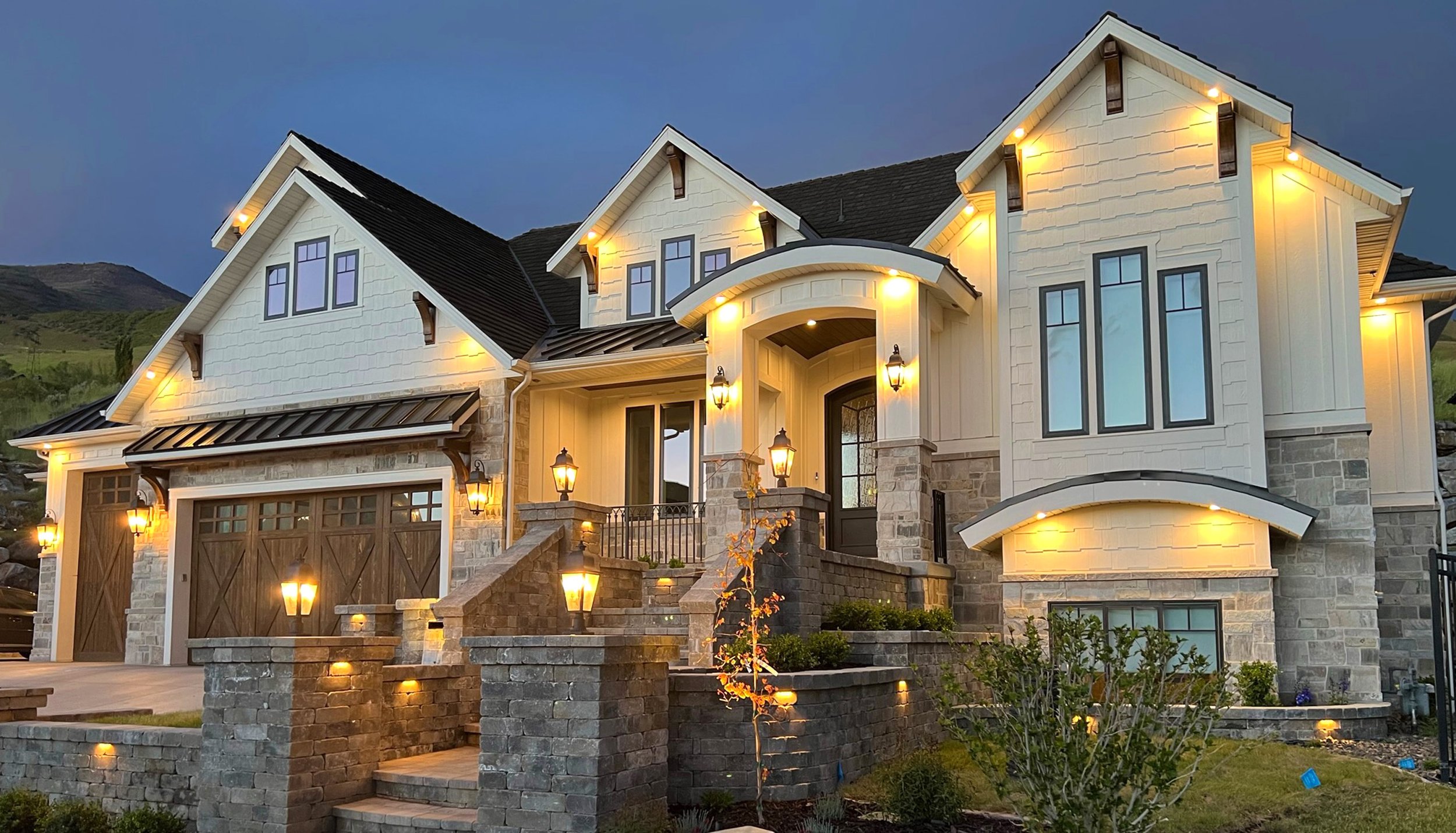 Exterior of a large, two-story house at dusk, illuminated by outdoor lights, with a stone and siding facade, multiple windows, a staircase leading to the front door, and a landscaped front yard.