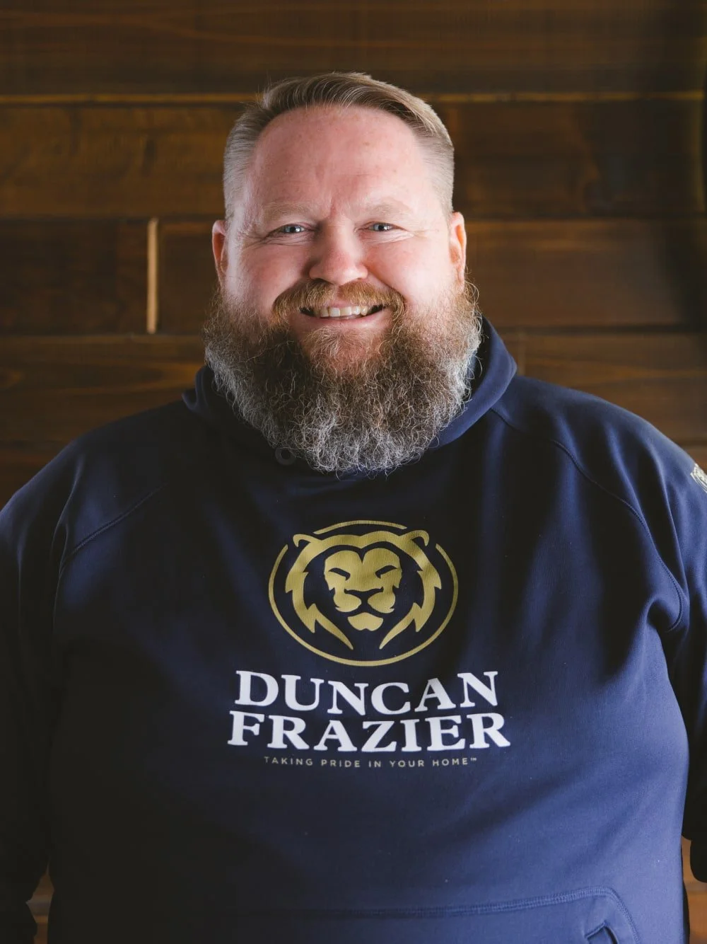 A smiling man with a beard and short hair wearing a navy hoodie with a gold lion logo and the text 'Duncan Frazier' and 'Taking Pride in Your Home' on it, standing in front of a wooden wall.