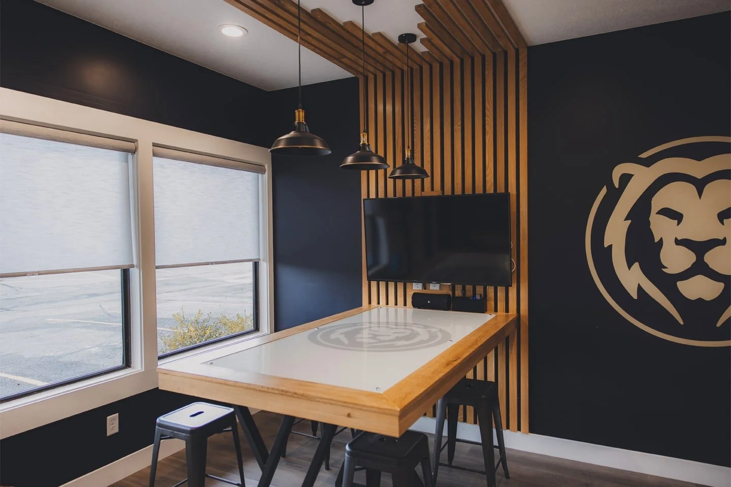 Modern conference room with wooden accents, a whiteboard table, black stools, a wall-mounted TV, large windows with blinds, and a lion logo on the dark wall.