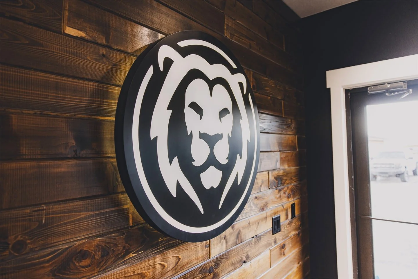 A black circular sign with a white lion's face logo mounted on a dark wooden wall near a door with a bright window.