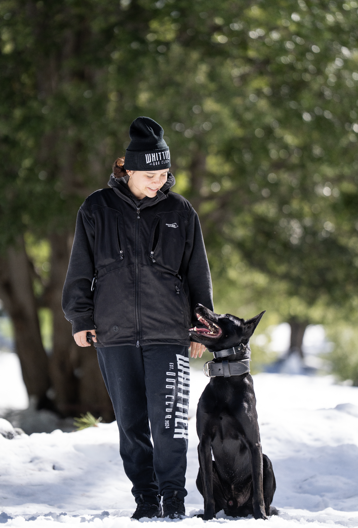 A woman in winter clothing with a black beanie and black jacket standing outdoors on snow, smiling at a black Belgian Malinois dog sitting beside her.