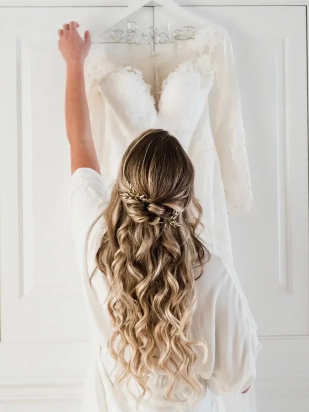 A bride with long, wavy blonde hair standing in front of a white door, reaching up to hang a glass wedding dress on a hook. The dress is white, with lace details, and a deep V neckline. The bride's hair is styled in loose curls with a braided section.