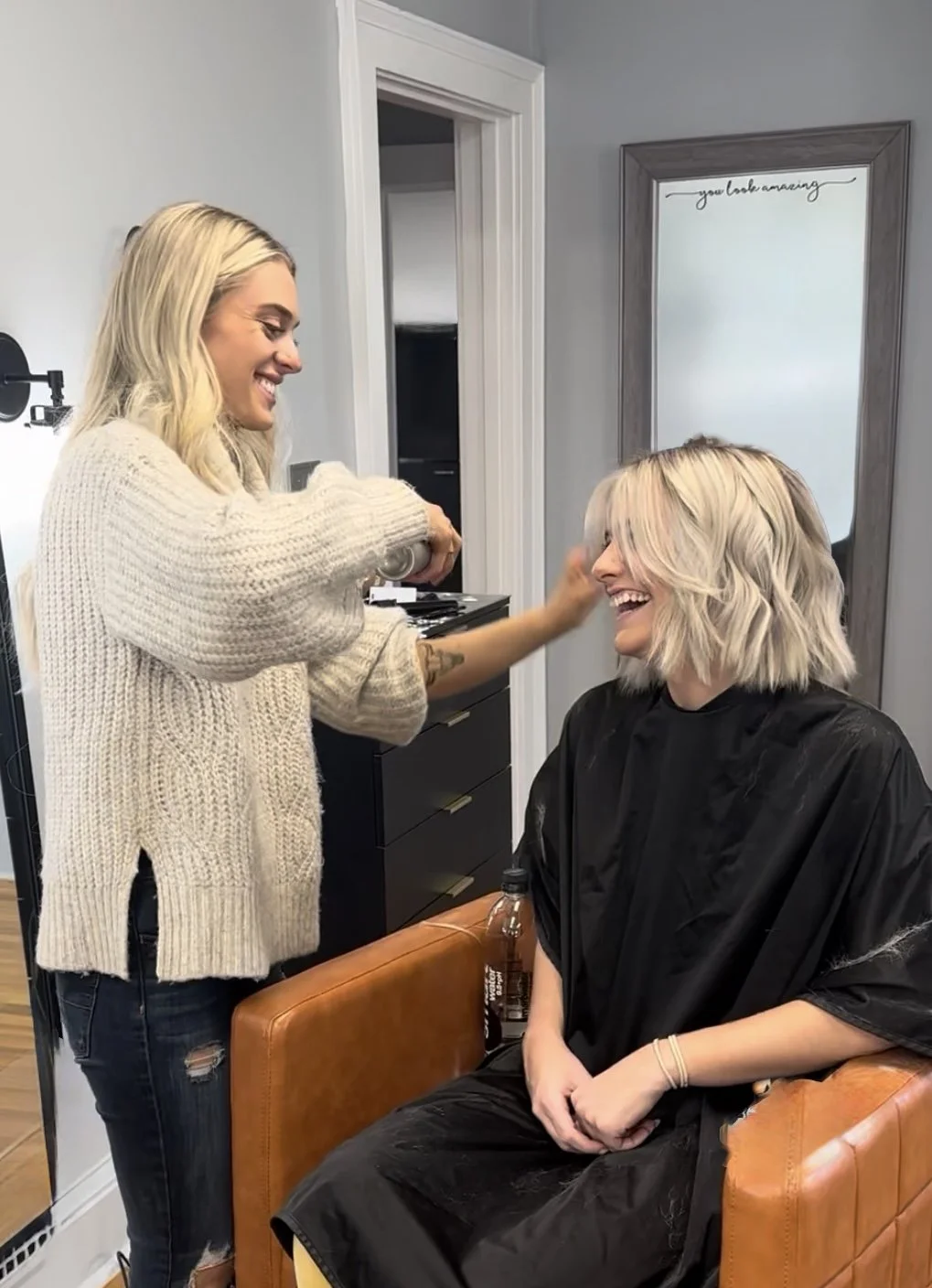 A hairstylist smiling and applying hair spray to a woman with platinum blonde, wavy hair at a salon. The woman is seated in a black cape in front of a mirror.
