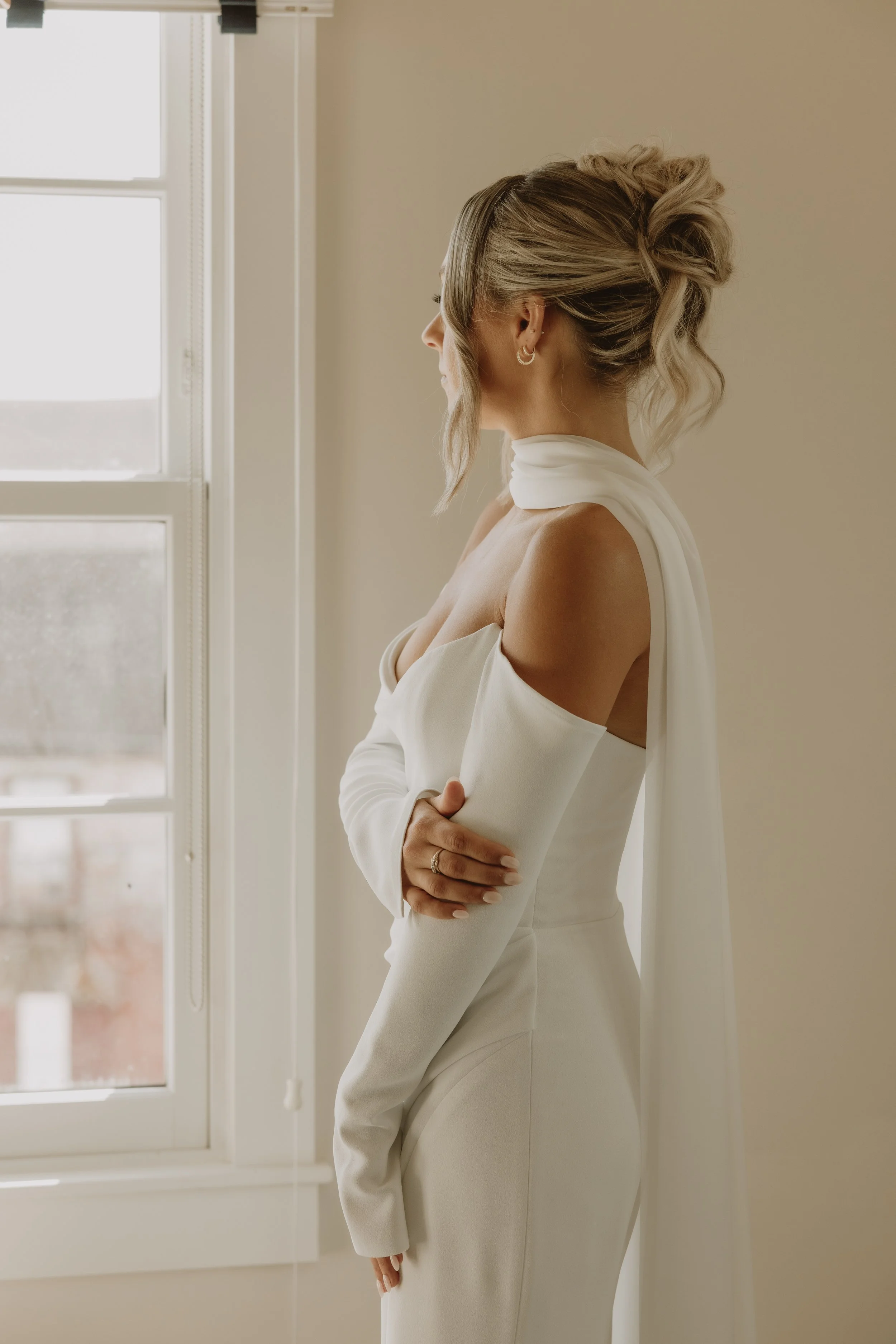 Woman in a white wedding dress with off-the-shoulder sleeves, standing near a window, looking to the side with her arms crossed.