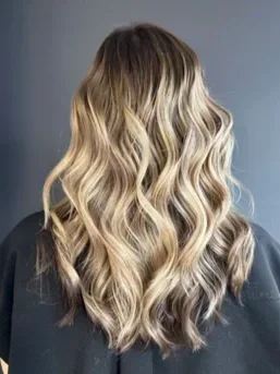 Back view of a woman with long, wavy blonde hair against a plain background.