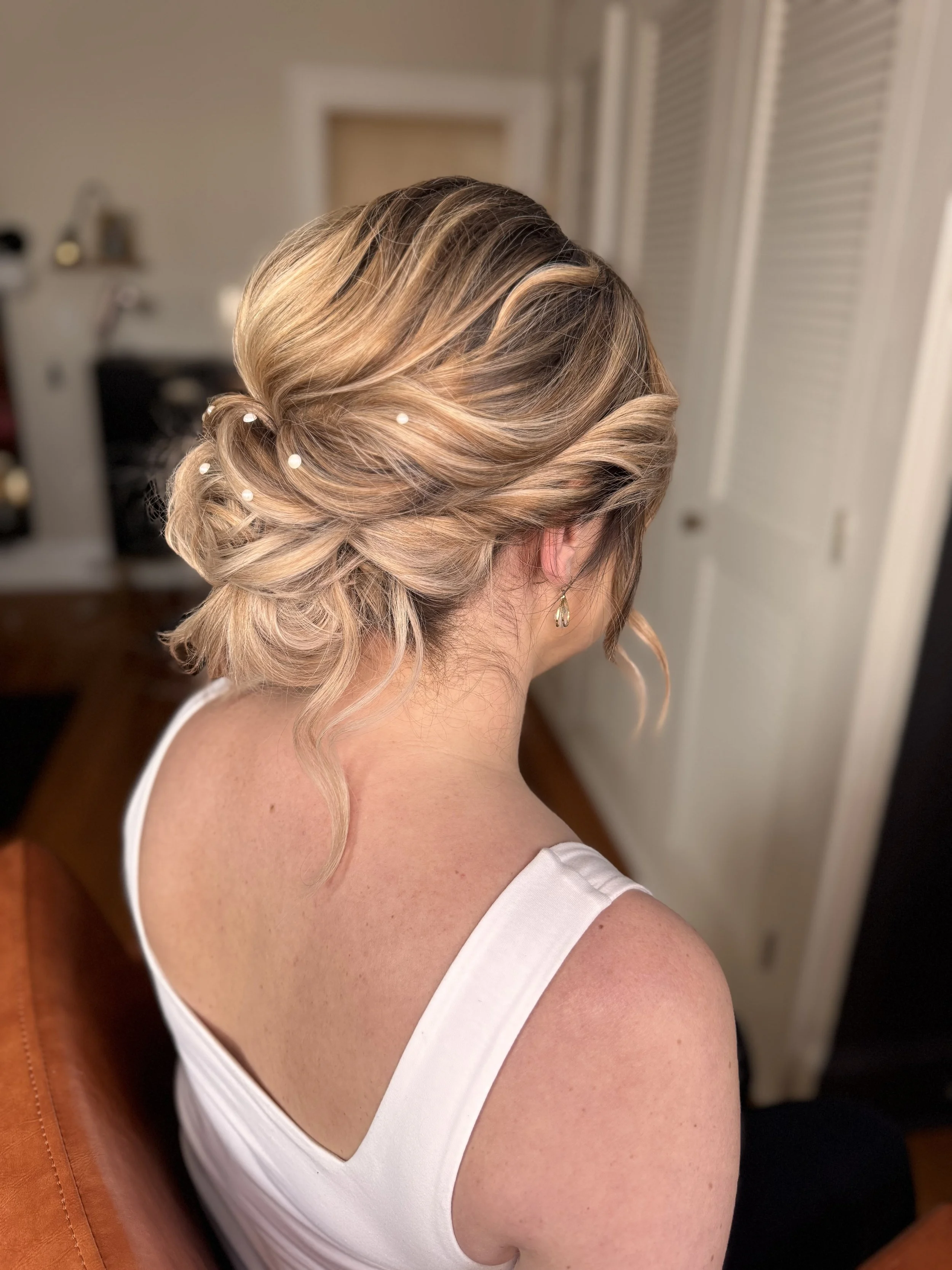 A woman with styled blonde hair decorated with small pearls, wearing a white sleeveless top, sitting in a room.