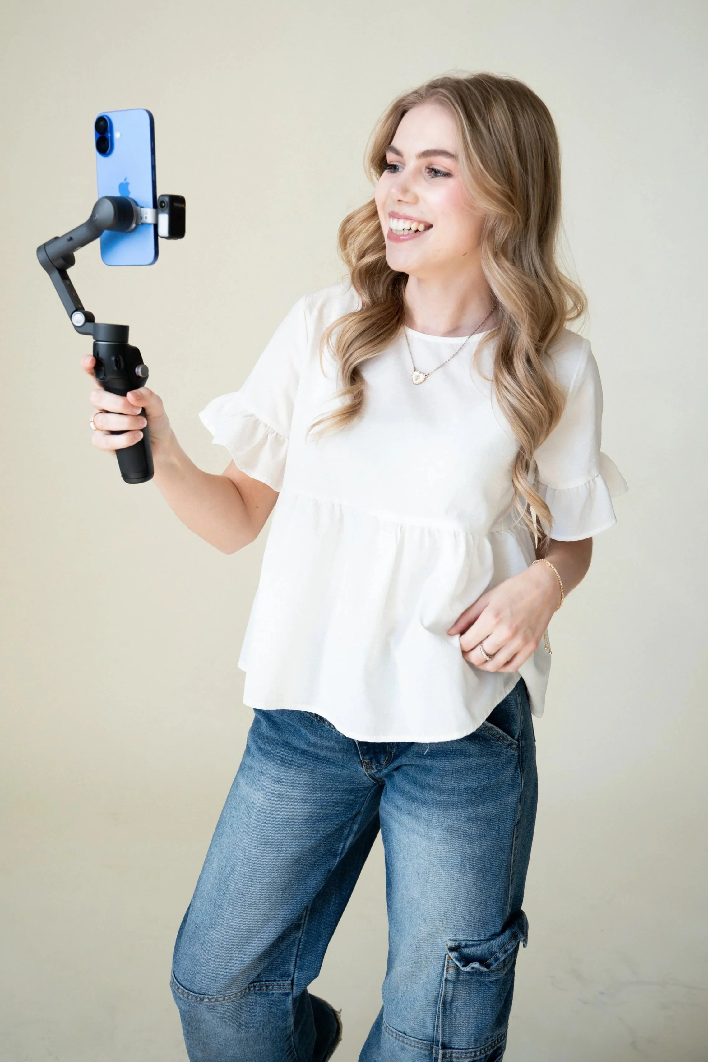 A woman with long, wavy hair wearing a white blouse and blue jeans is holding a smartphone mounted on a handheld stabilizer, taking a selfie and smiling.