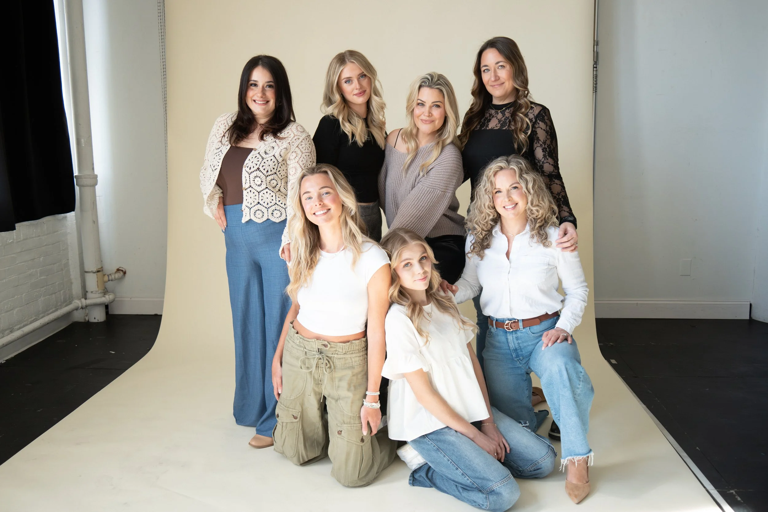 A group of nine women of various ages and blonde, brunette and red hair, posed together against a beige backdrop in a photo studio.