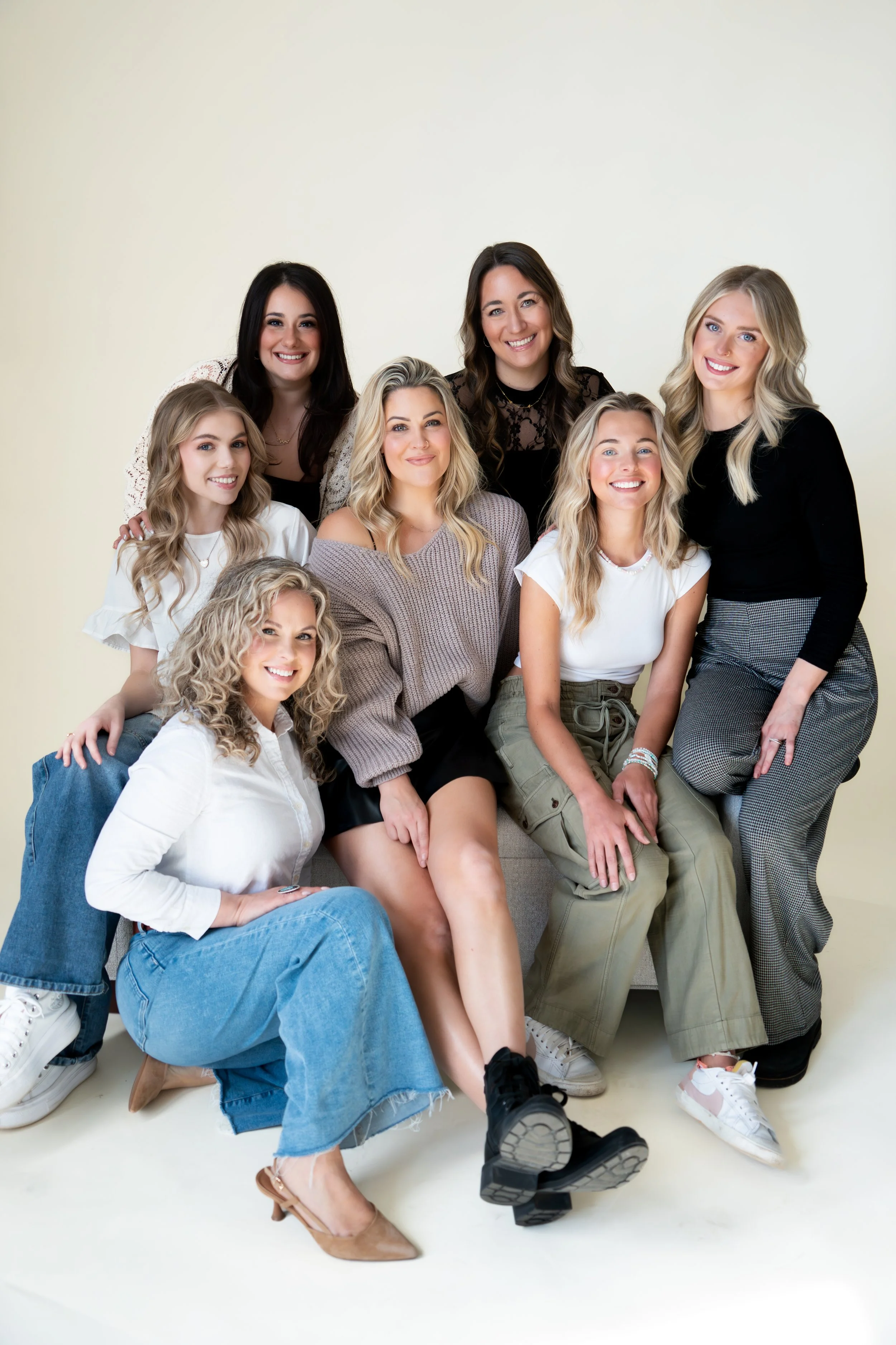 Group of eight women smiling and sitting together inside in front of a plain light background.