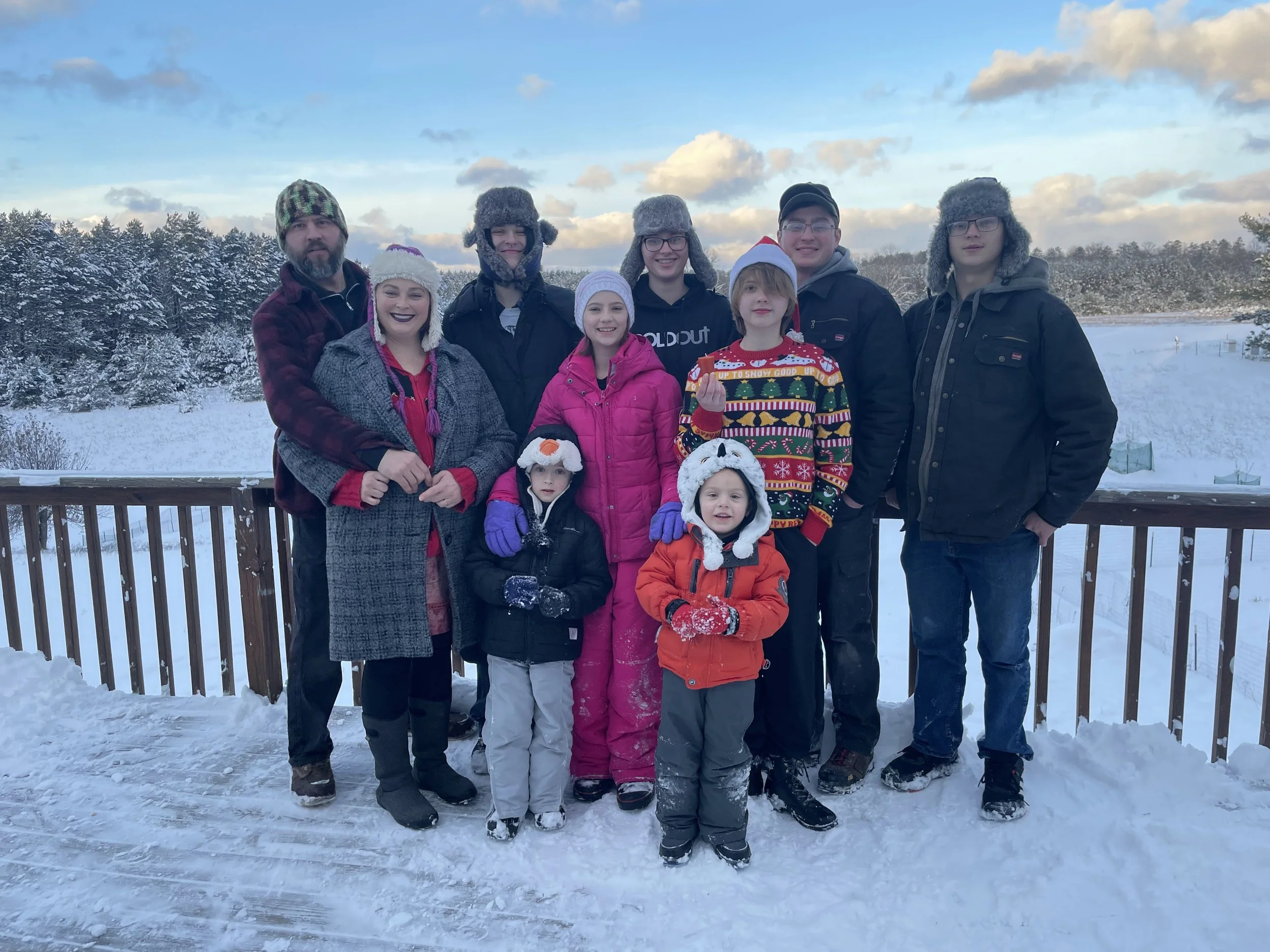 Family group of ten people, including children and adults, dressed warmly in winter clothes and standing on a snow-covered deck outdoors with a snowy landscape and partly cloudy sky in the background.