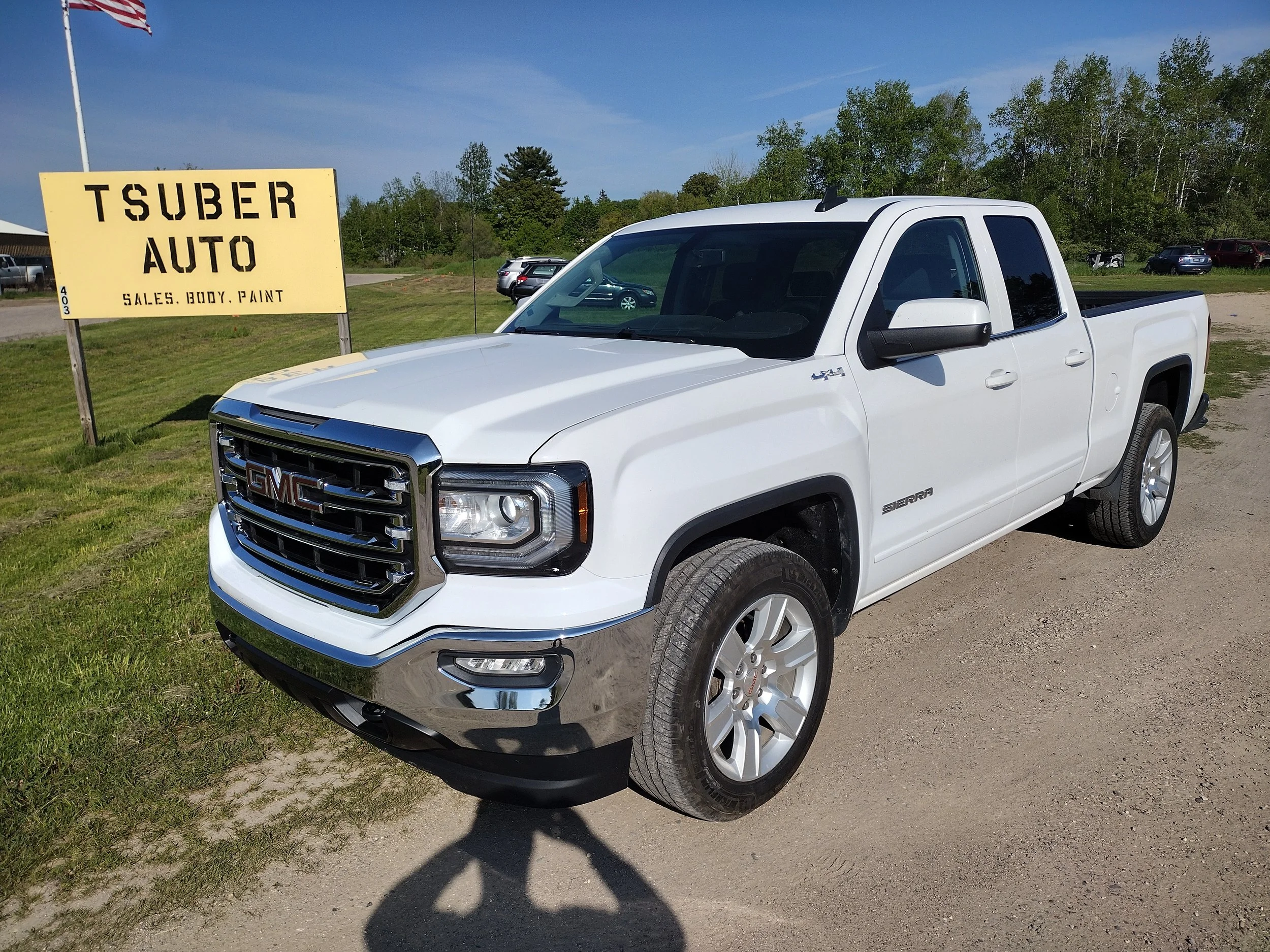 White GMC Sierra pickup truck parked outside on a dirt surface with a Tsuber Auto sign in the background advertising sales, body, and paint services.