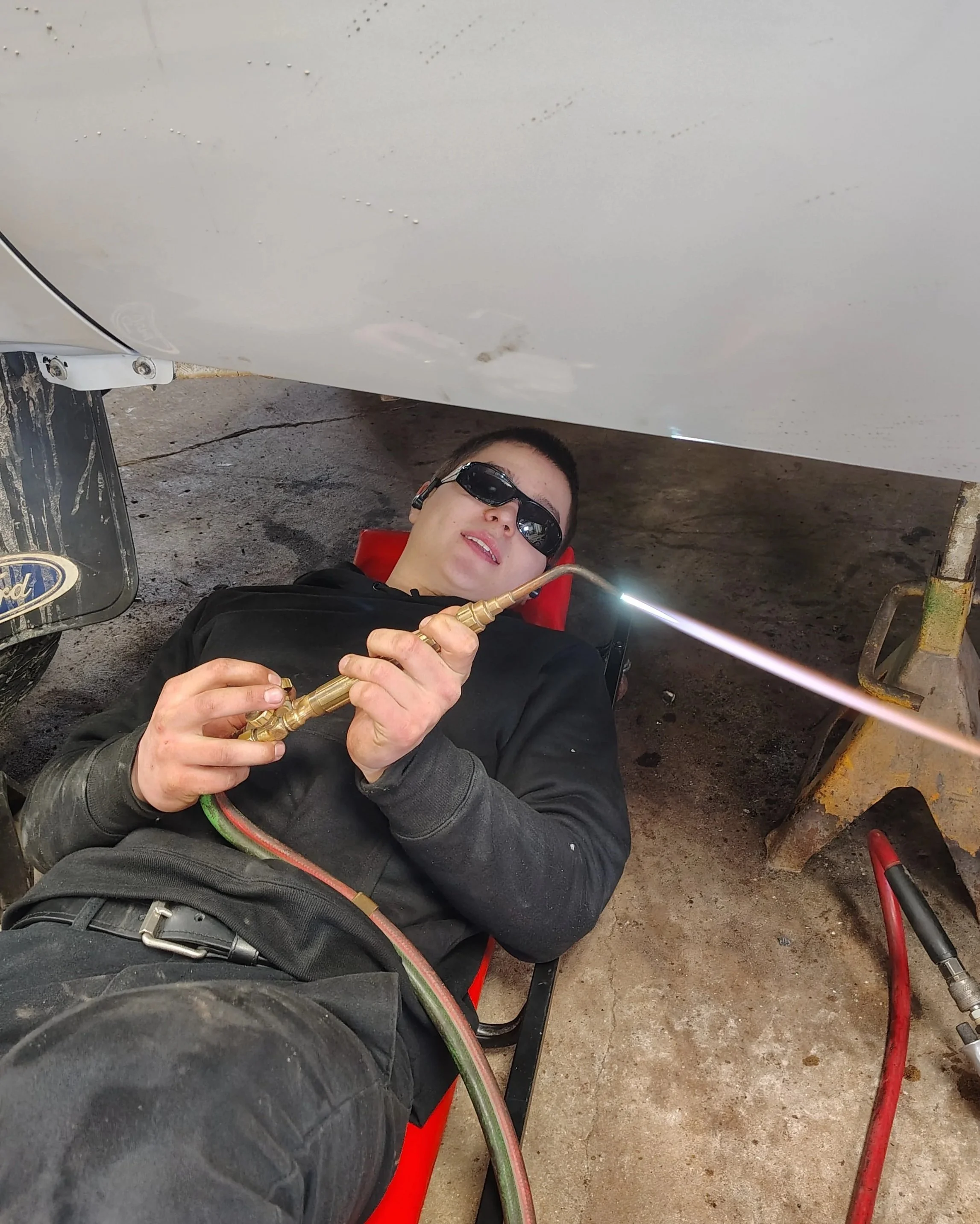 A man wearing sunglasses and black clothing is lying on a creeper under a vehicle, welding with a torch. There are tools and equipment around him, and the ground is concrete.