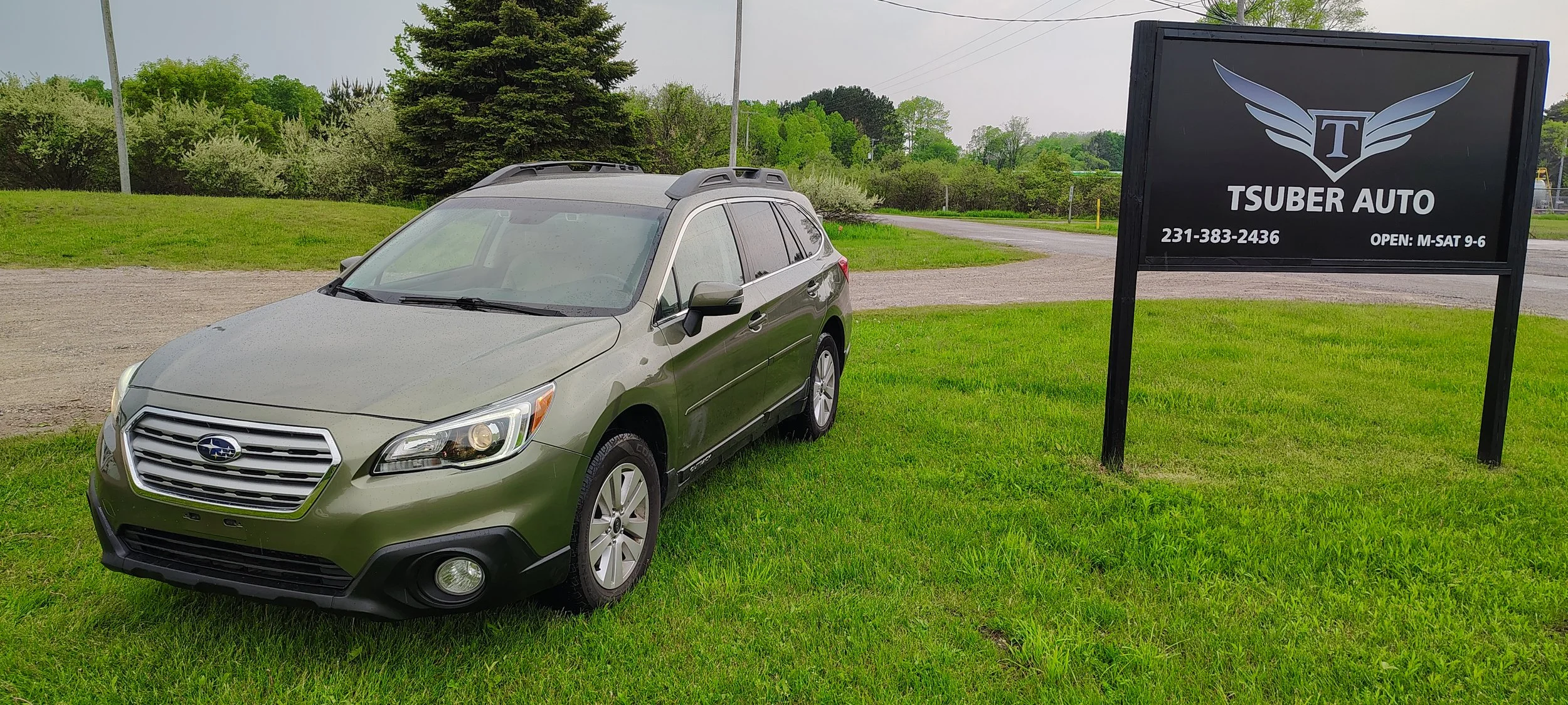 A green Subaru car parked on a grassy area next to a black sign for Tsuber Auto with contact information and business hours, in a rural setting with trees and a cloudy sky.
