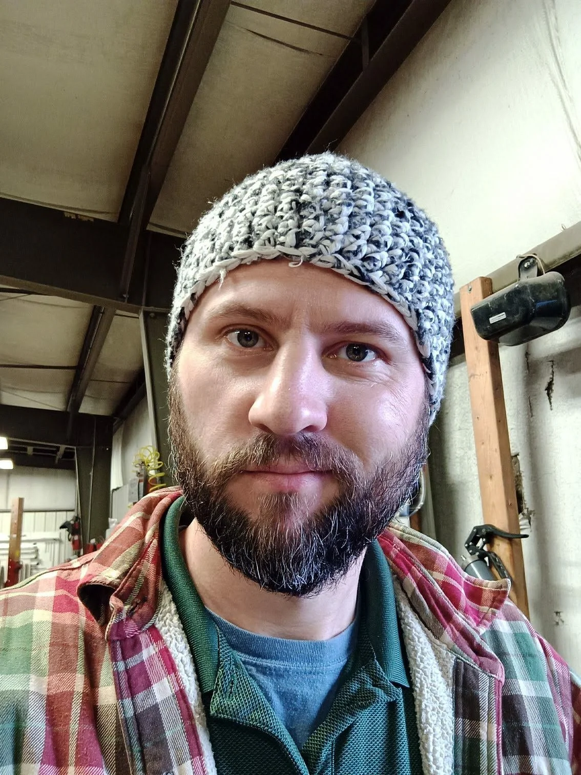 A man with a beard wearing a gray knit hat and a plaid shirt taking a selfie inside a workshop or garage.