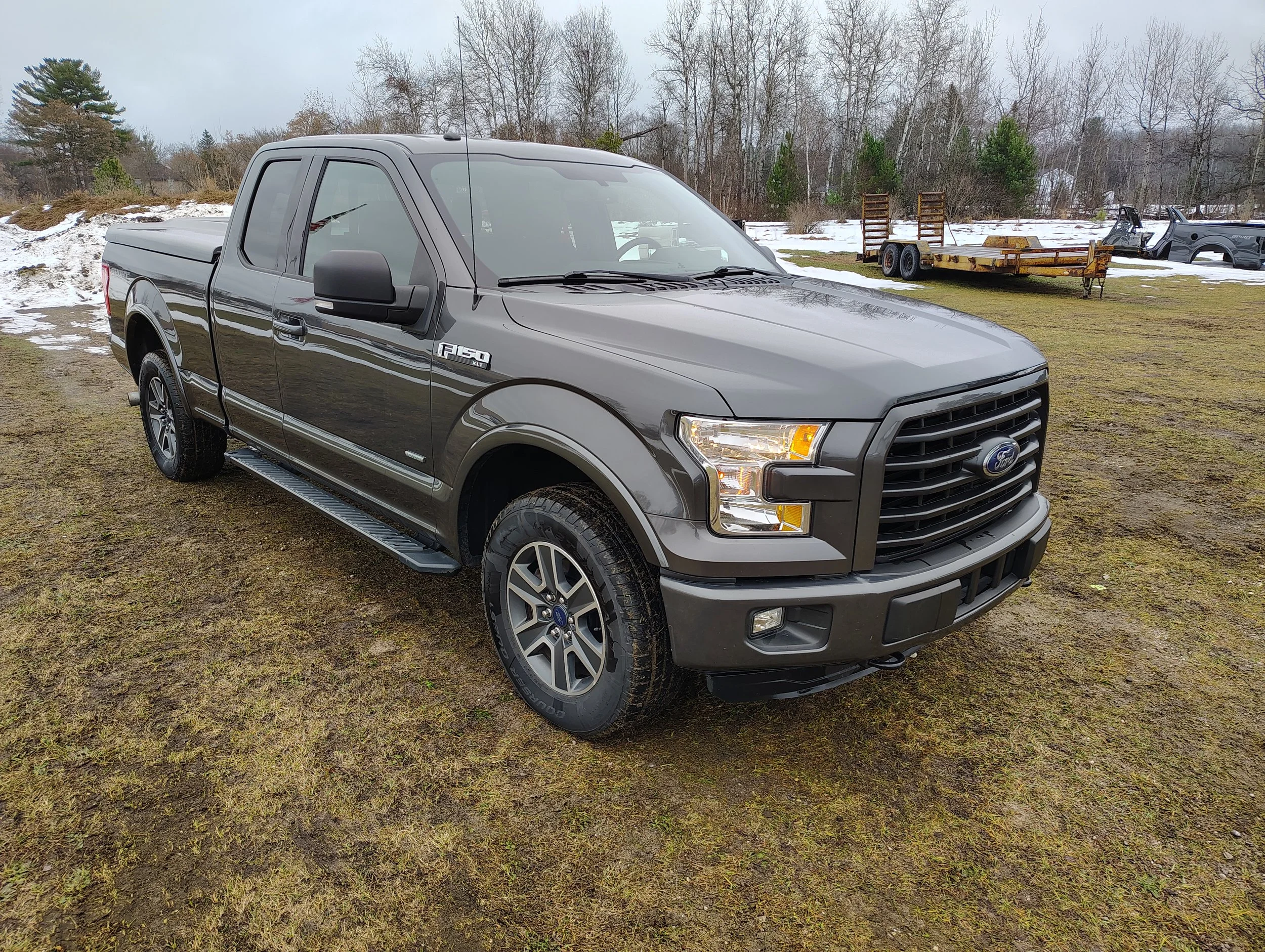Grey Ford F-150 pickup truck parked on grassy ground with patches of snow, trees, and trailers in the background.