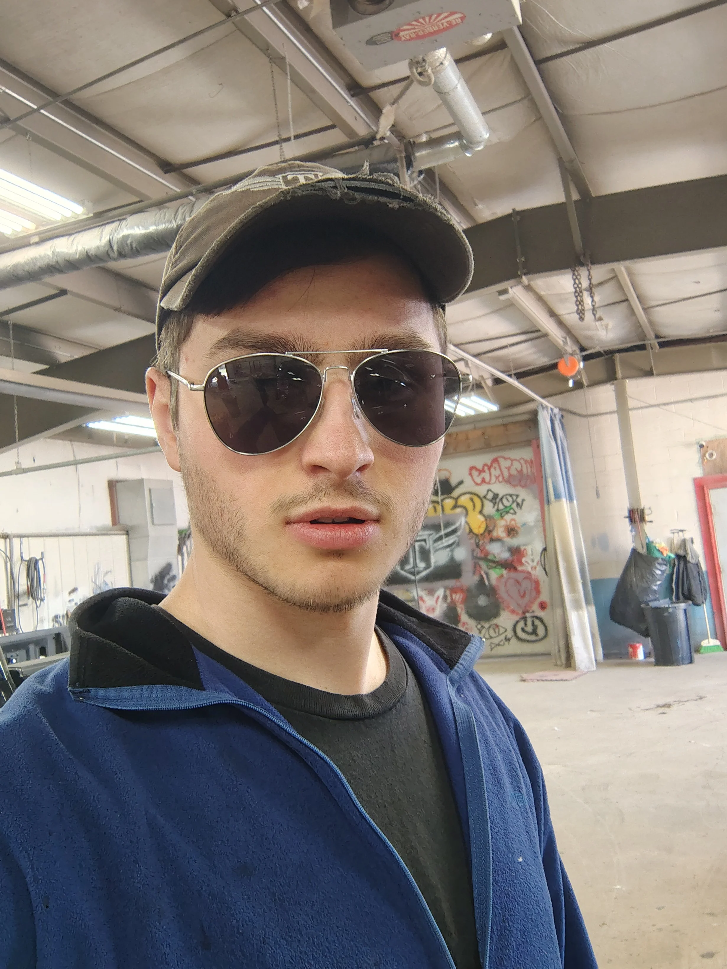 A young man with short brown hair and light skin wearing dark sunglasses, a camouflage baseball cap, and a blue zip-up jacket in an industrial warehouse with graffiti on the wall.