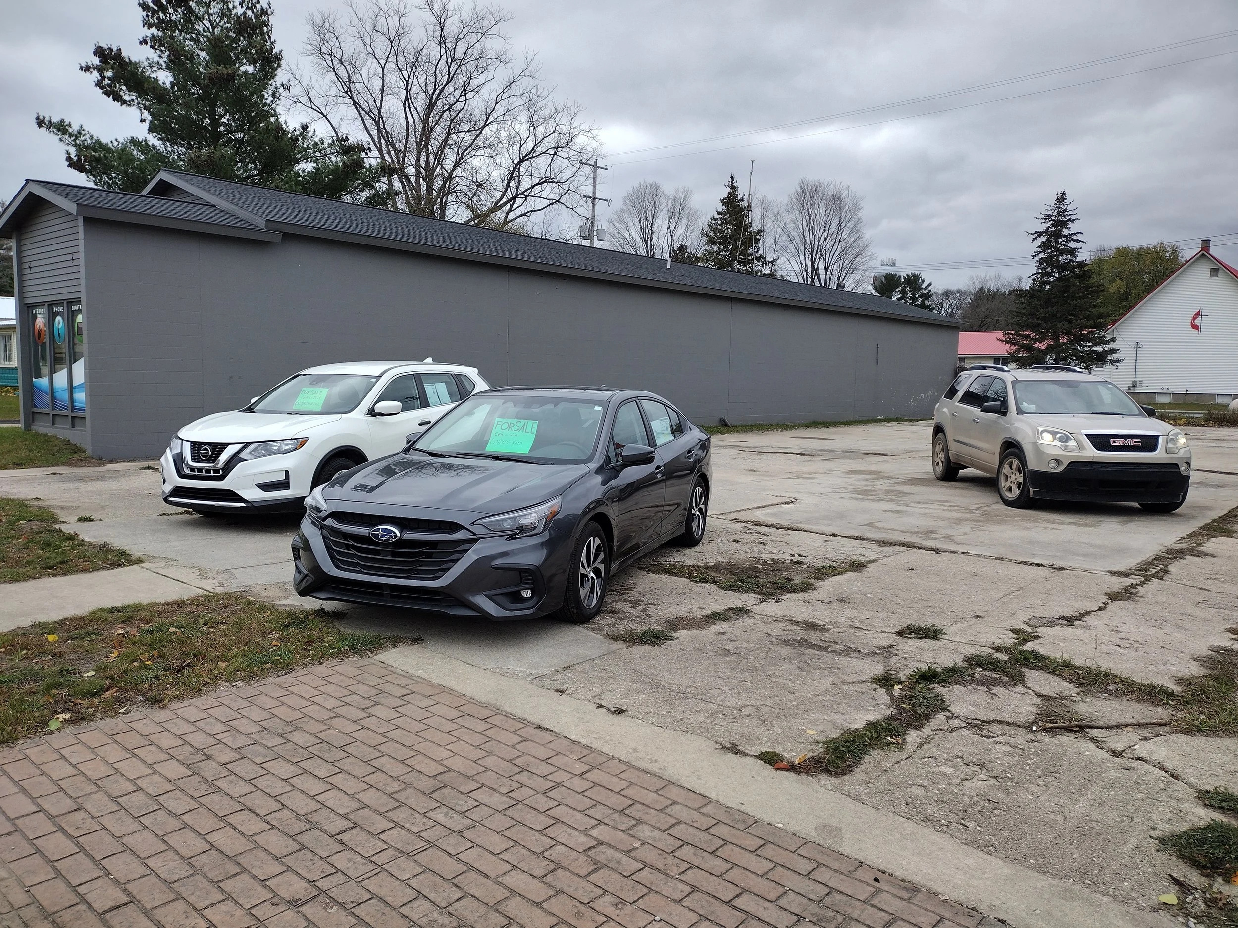 Three cars parked on a cracked parking lot in front of a gray building with a blue storefront to the left. The car in the middle has a "For Sale" sign, and the one on the left has a "Sale" sign in the window. There are trees and a house in the background under a cloudy sky.