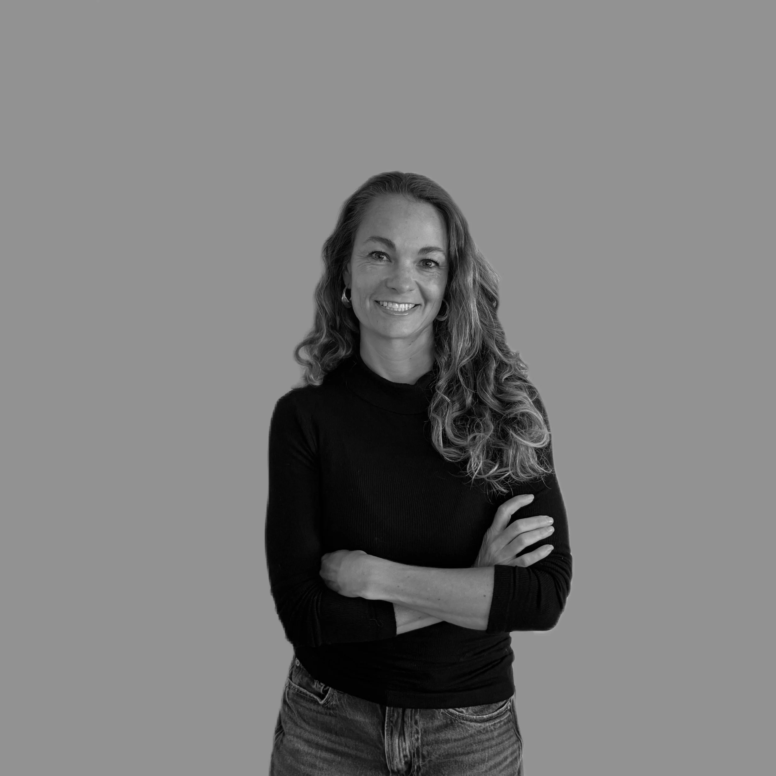 A woman with curly hair and hoop earrings smiling, wearing a black long-sleeve shirt and jeans, standing against a gray background.