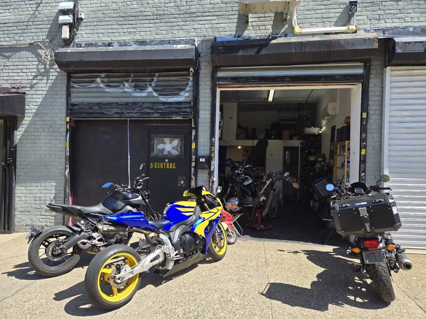 An open garage with motorcycles parked outside and tools inside, adjacent to a brick building with a metal roll-up door.