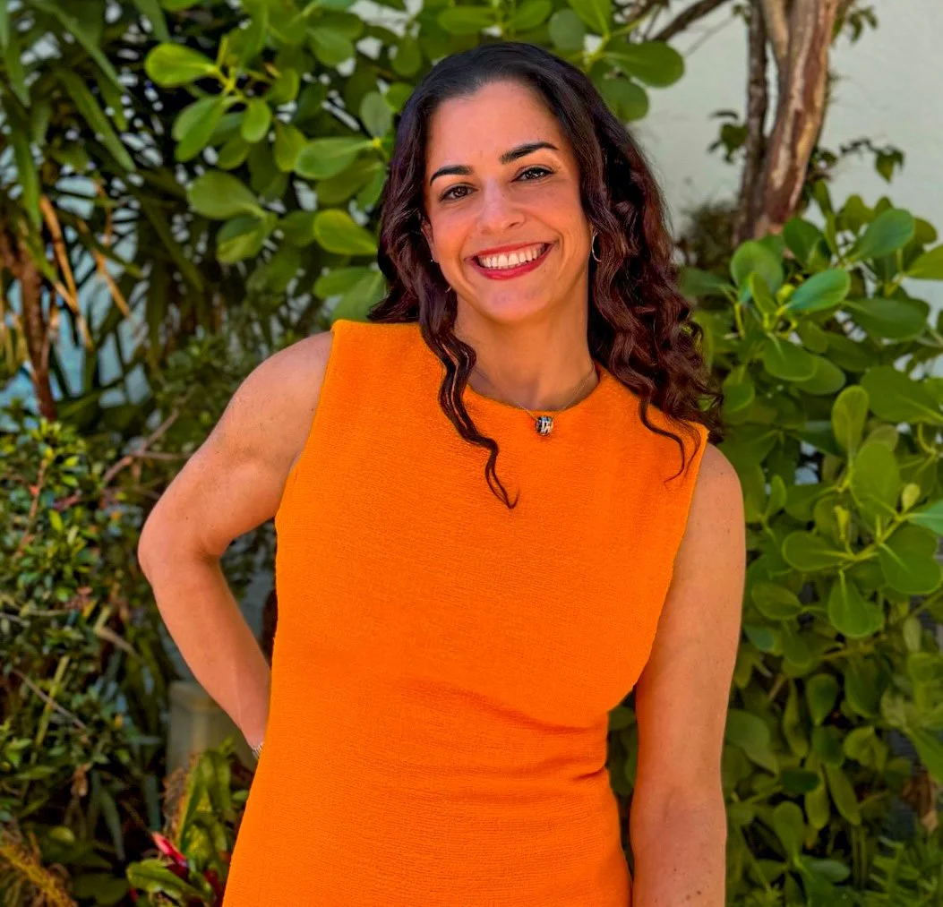 A woman with dark curly hair smiling outdoors in front of green leafy plants, wearing an orange sleeveless dress and a necklace.